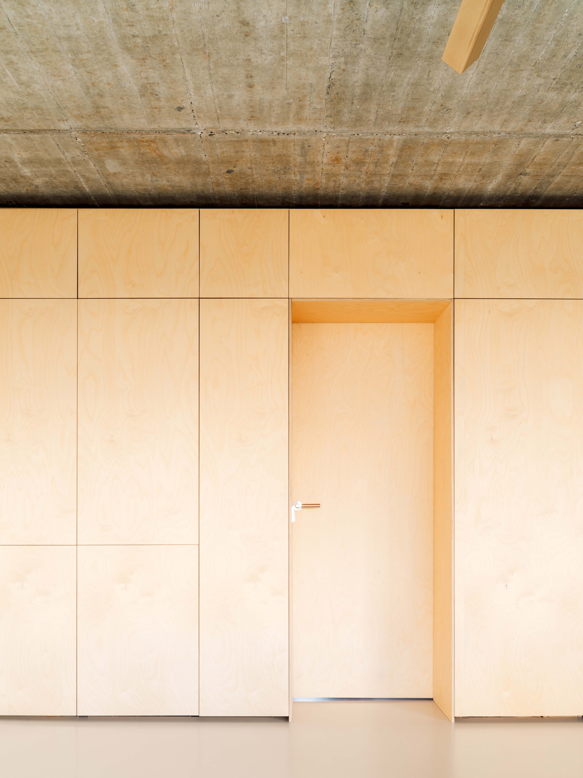 Straight-on view of the birch plywood wall with a flush door and handle below a board-marked concrete ceiling