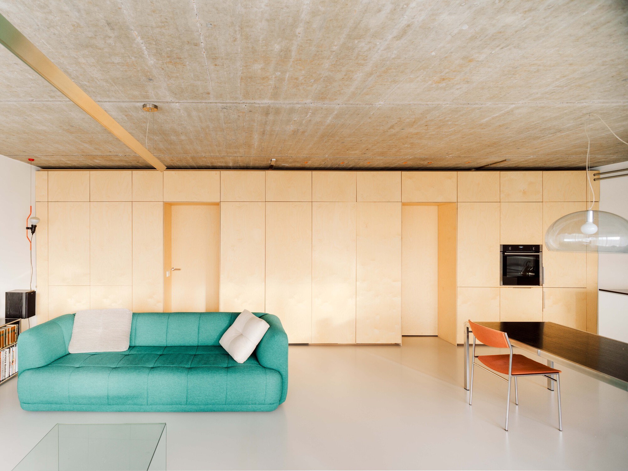 Living area with a turquoise sofa in front of a birch plywood storage wall, under a board-marked concrete ceiling in daylight