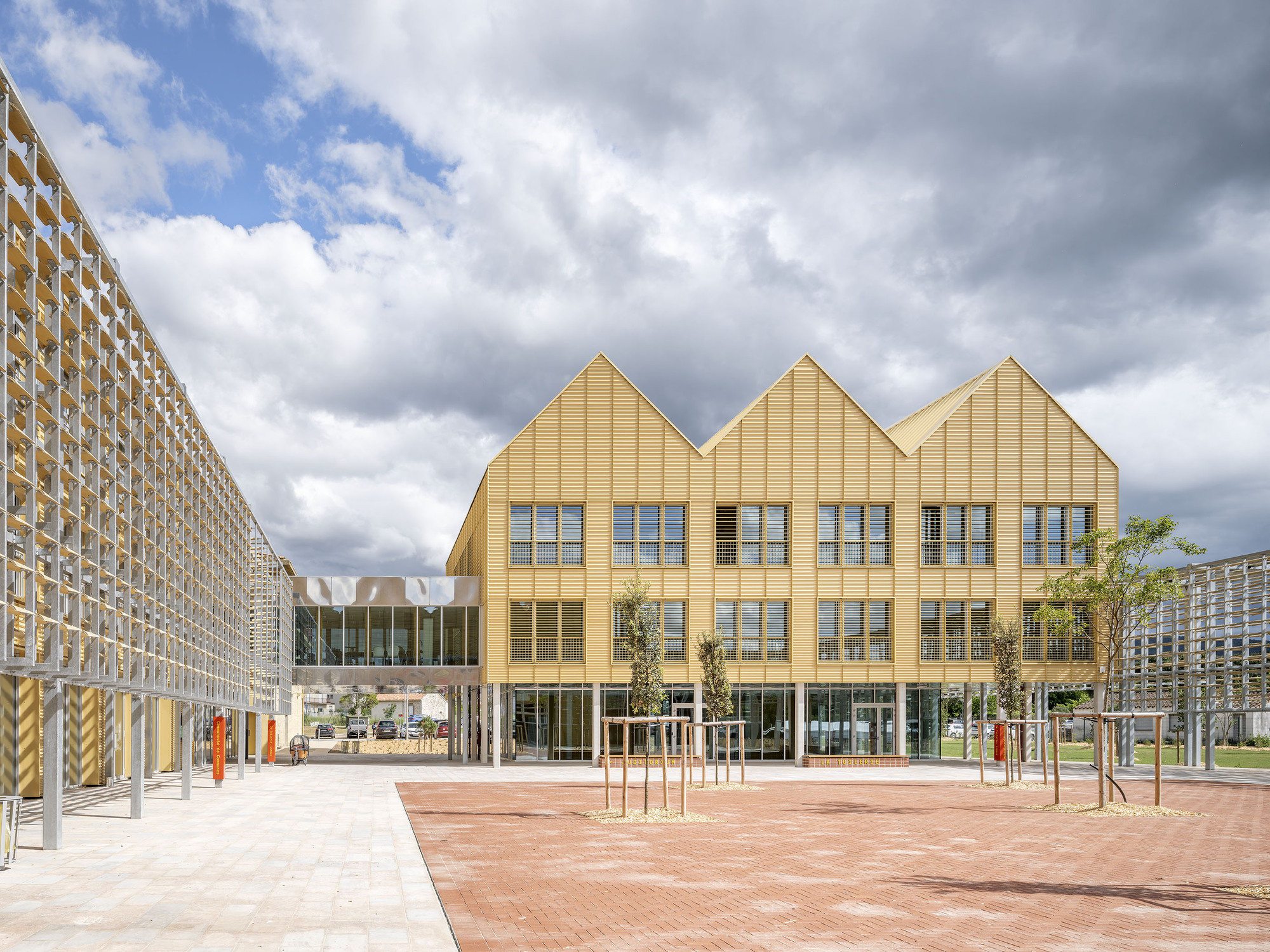 Three-gabled yellow ribbed facade across a brick paved square, with a lattice screen at left under heavy clouds