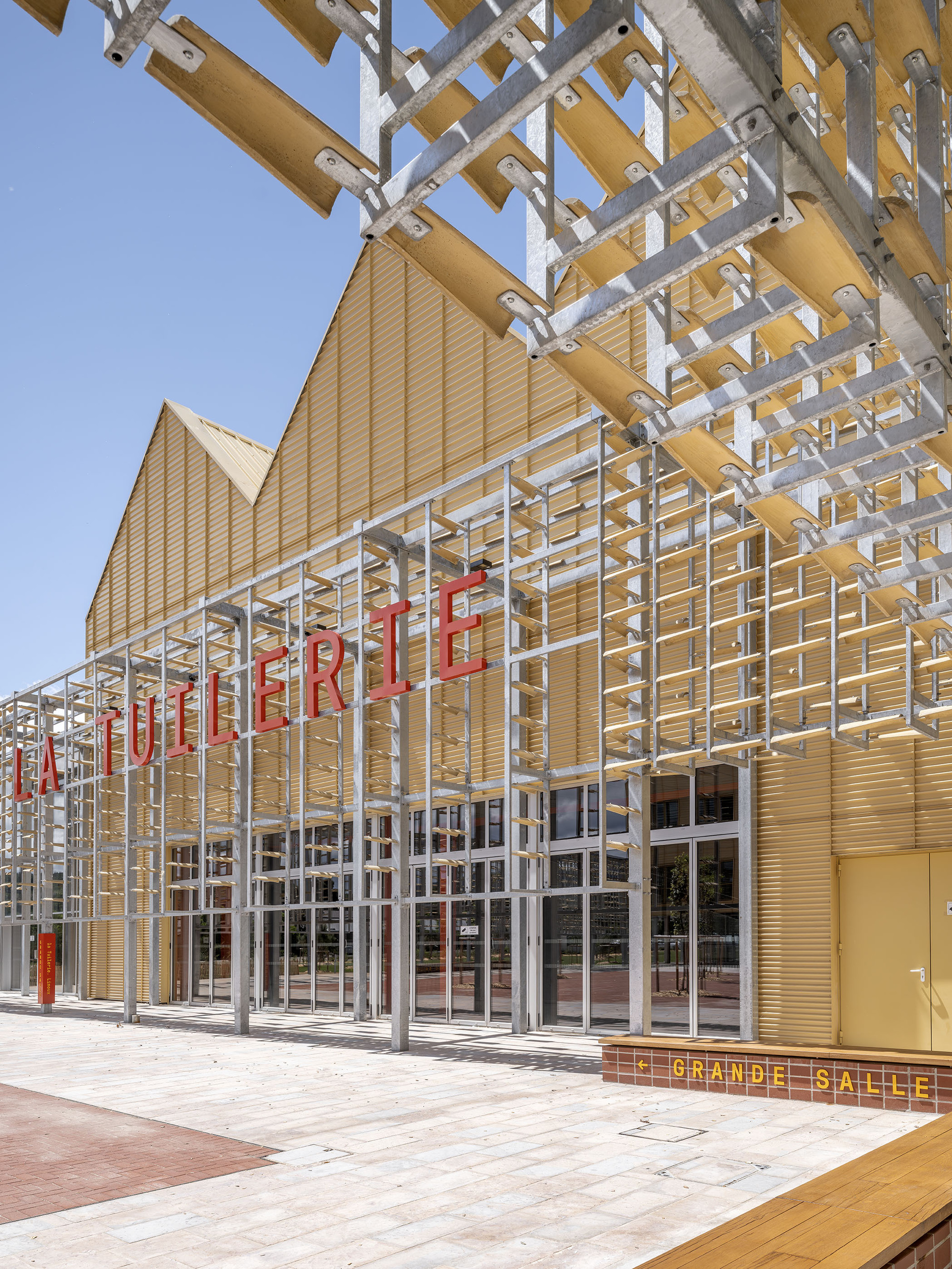 Galvanized steel frame with timber slats shading a glazed entrance, red lettering above a bench in clear daylight