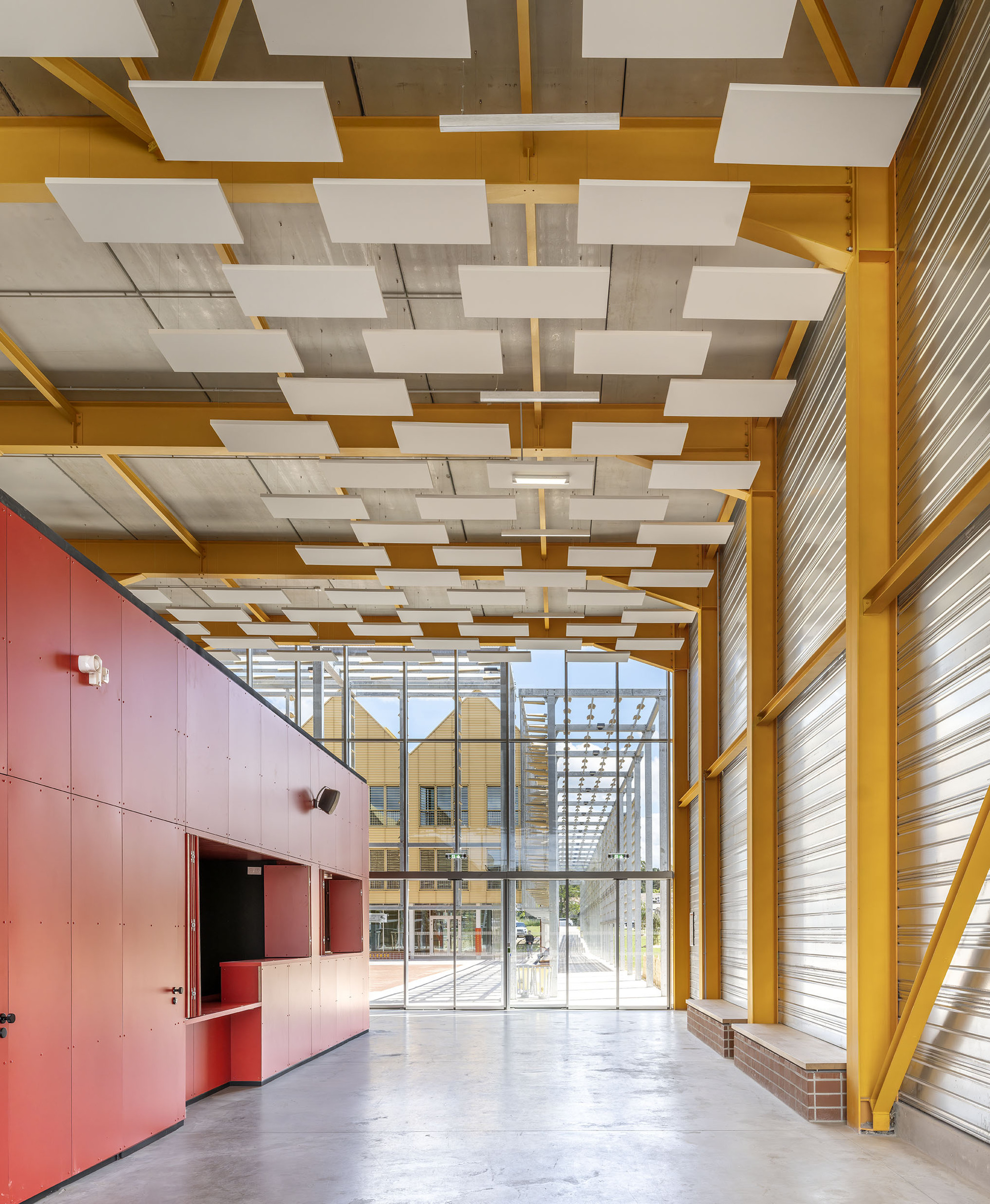 Double-height hall with yellow steel frame, red cabinet wall and white acoustic ceiling panels, glazed end opening to the square