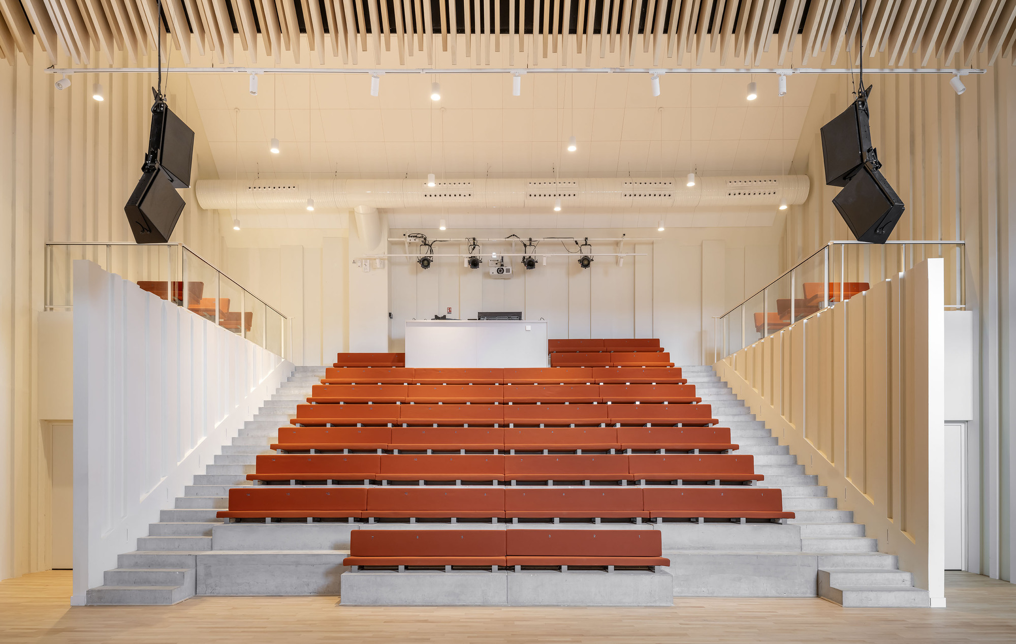 Tiered auditorium with red benches on concrete steps, timber slat walls and ceiling, and speakers hung either side
