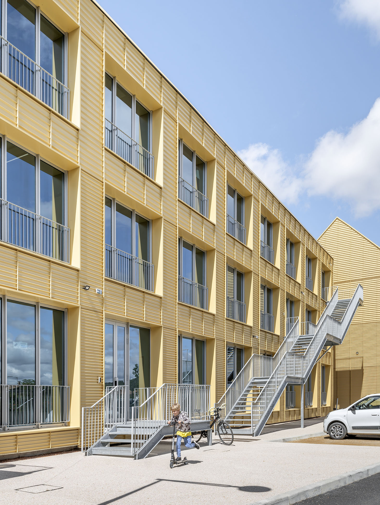 Yellow corrugated facade with recessed balcony windows and steel exterior stairs, a child on a scooter below a parked car