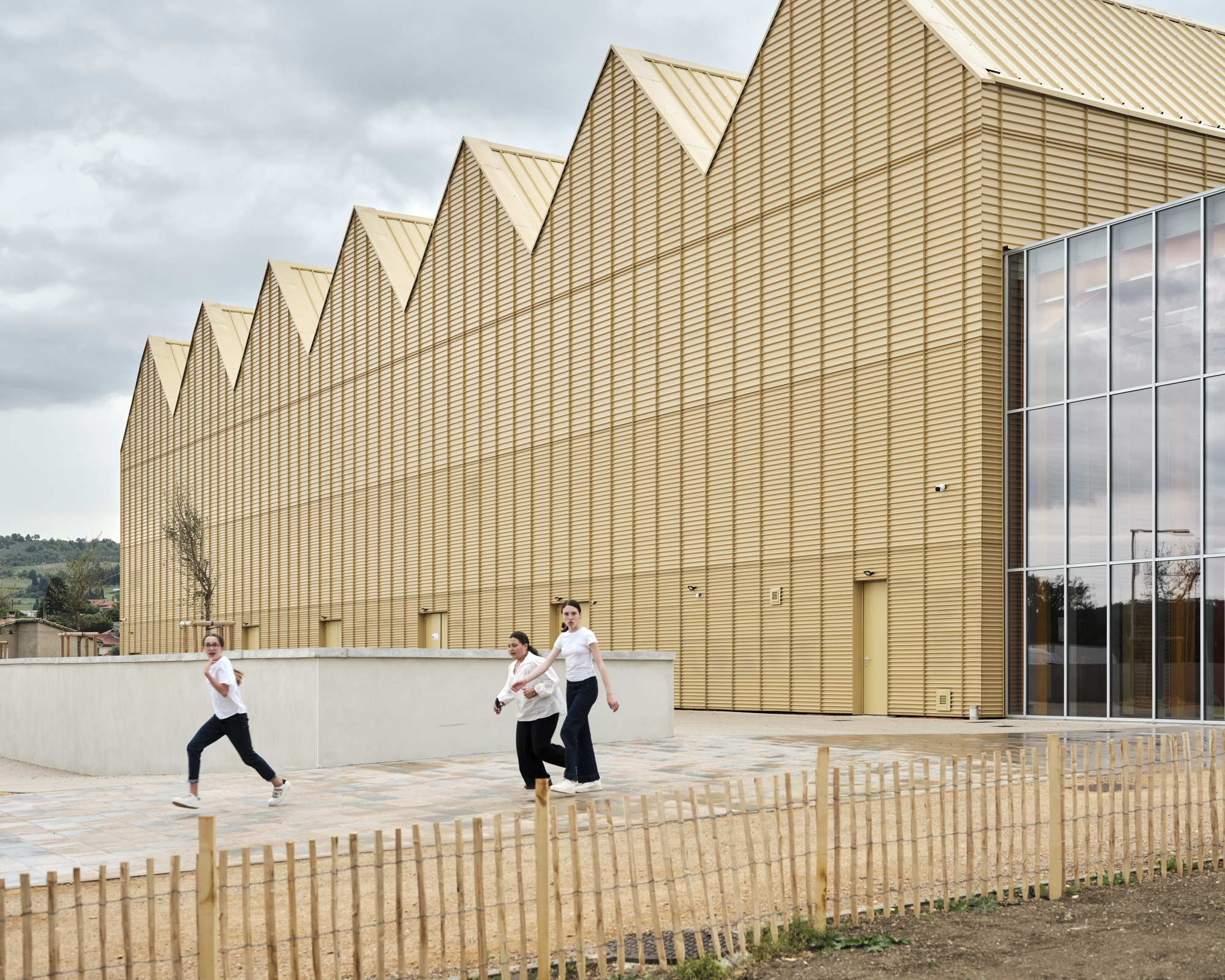 Sawtooth gable facade clad in gold corrugated metal, with three people running past a low concrete wall under an overcast sky