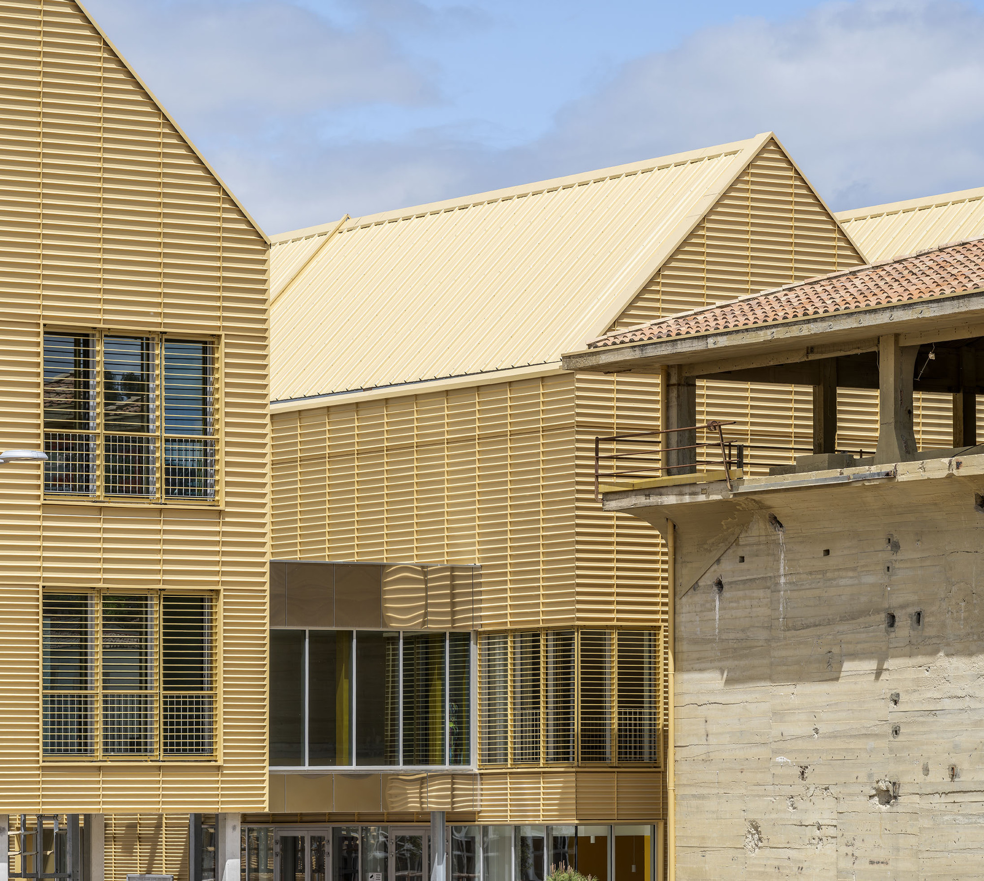 Gabled gold louvered volumes beside a raw board-marked concrete structure with a terracotta tile roof, under a blue sky with clouds