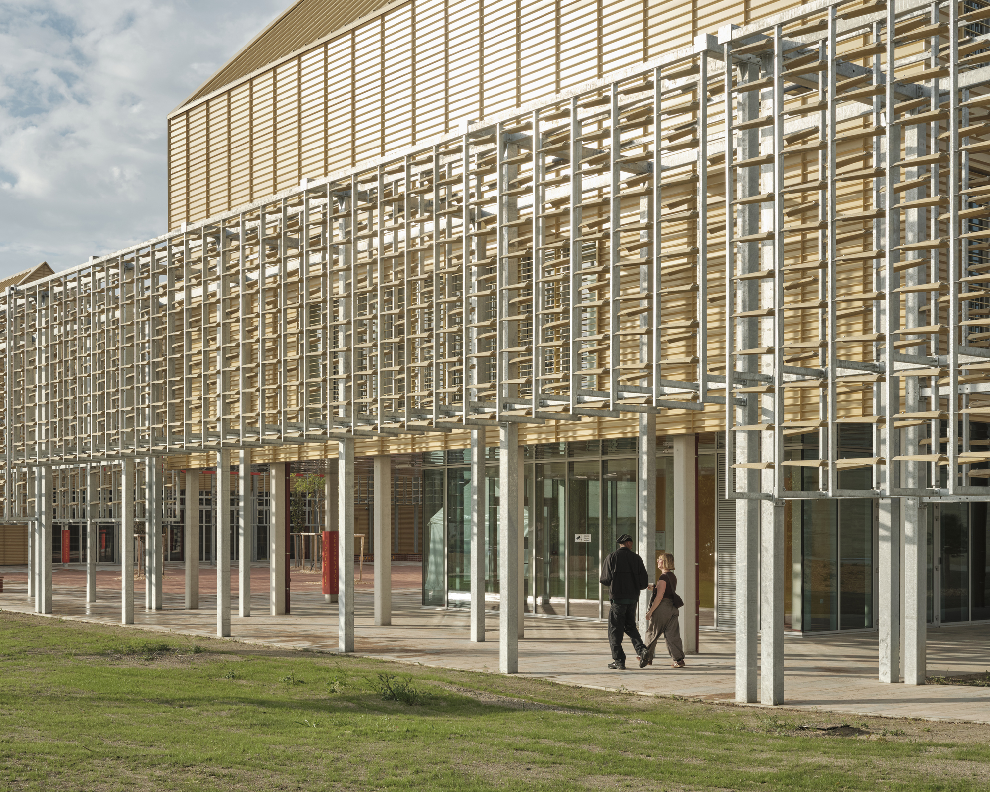 Galvanized steel trellis screen on slender columns in front of the gold facade, with two people walking across the paving