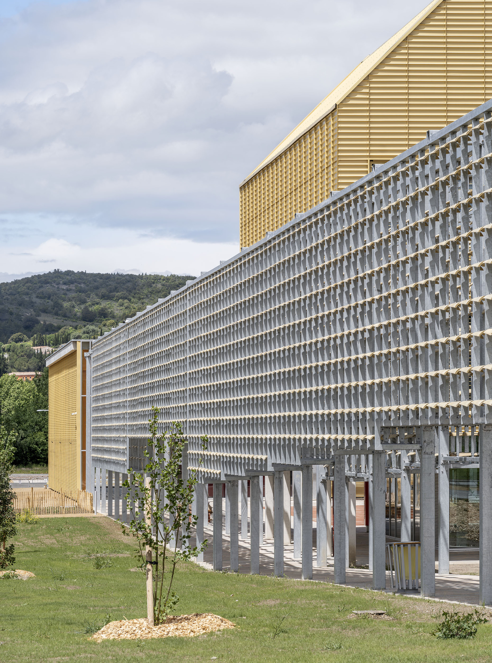 Long perforated grey screen wall raised on thin columns, with a young tree and wooded hills behind under cloudy skies