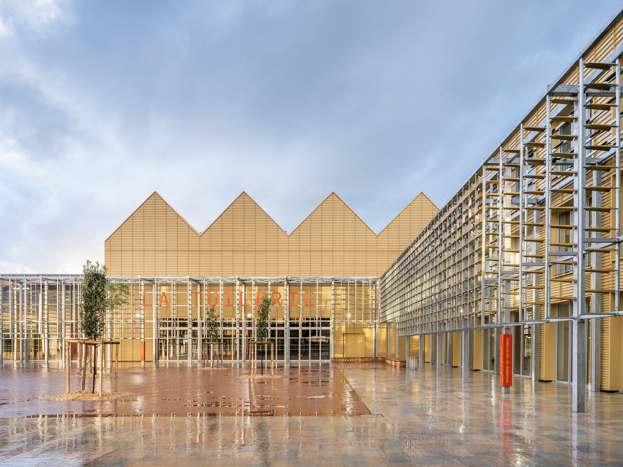 Courtyard of wet paving reflecting the sawtooth gold roofline and the steel screen, with saplings under a stormy sky
