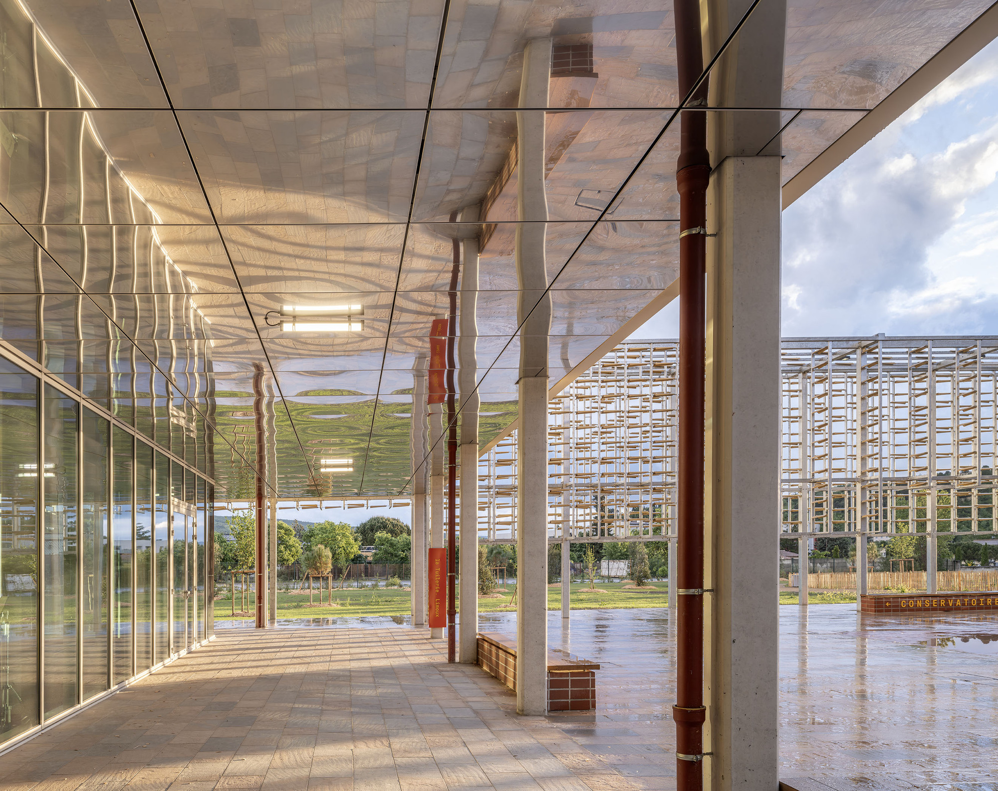 Covered walkway with reflective mirrored soffit, concrete columns and a copper-red downpipe, facing a timber lattice structure across a rain-wet plaza