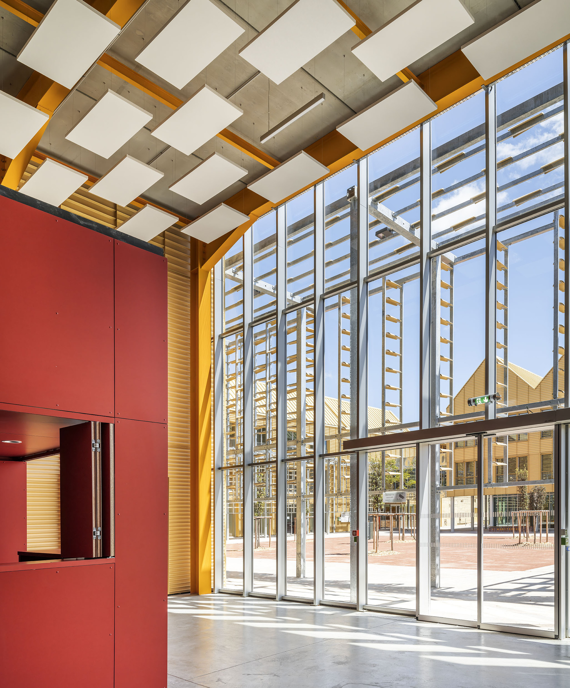 Double-height glazed lobby with yellow timber frame, red cabinet wall and floating white acoustic panels, sunlight casting shadows on the concrete floor