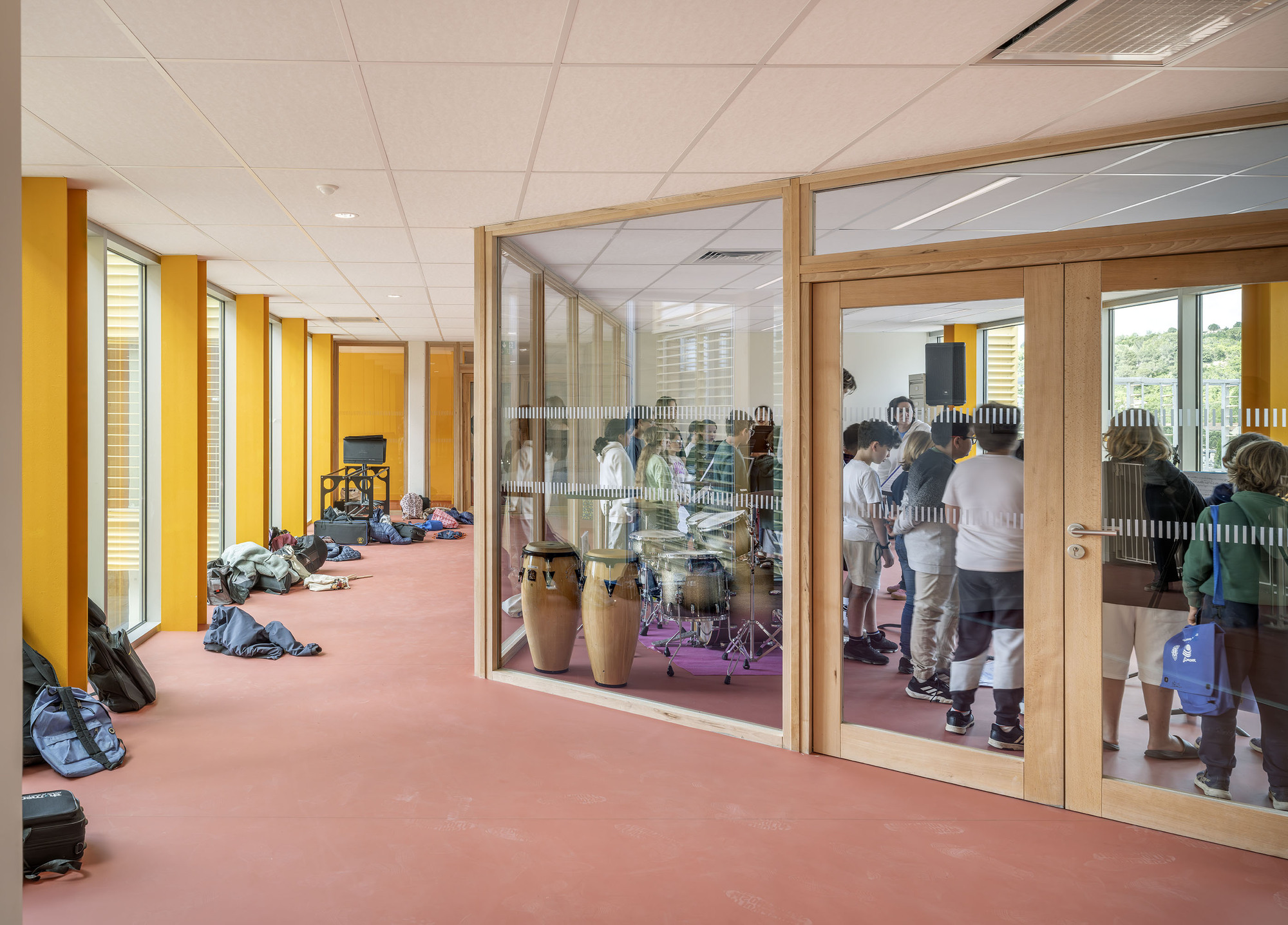 Corridor with yellow columns and red floor beside a glazed timber-framed music room where students gather around drums