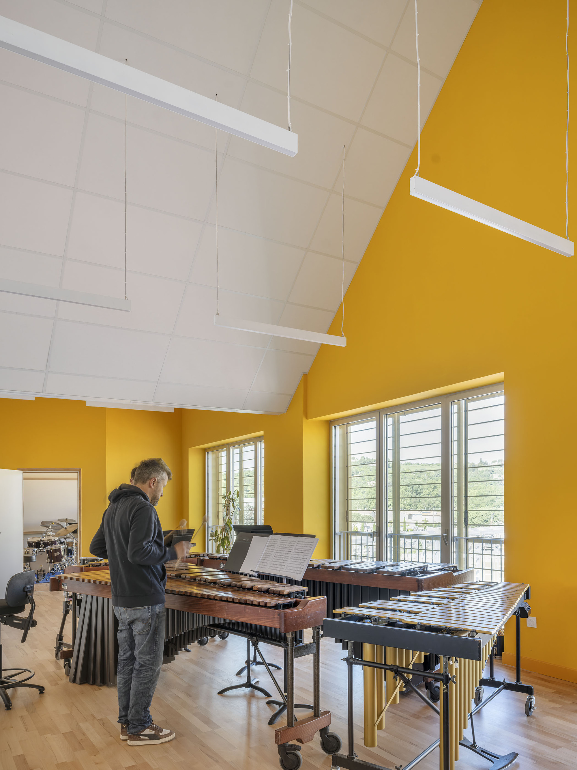 Music room with steep yellow walls and pitched white ceiling, a man standing at a marimba in daylight from the windows
