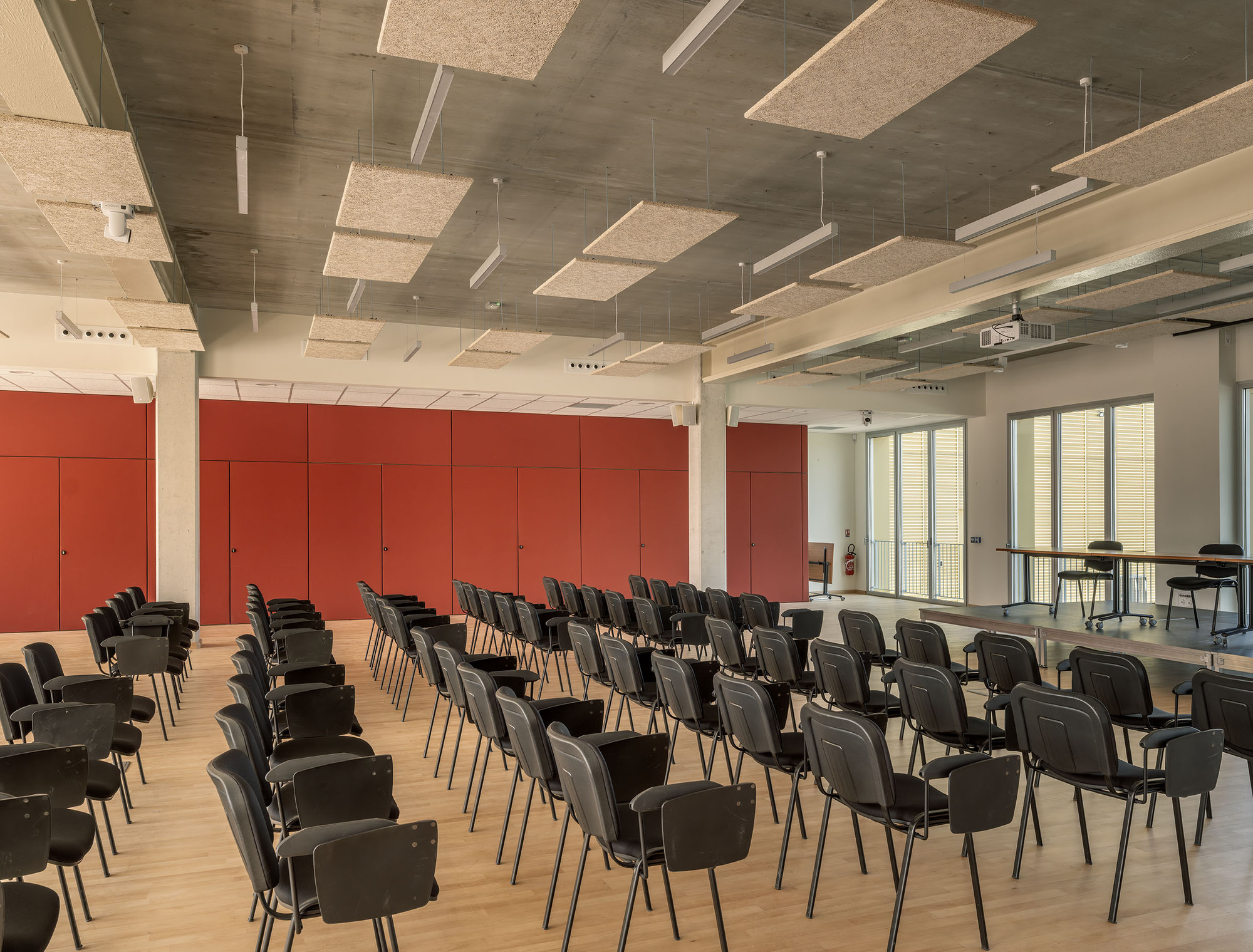 Multipurpose hall with rows of black chairs facing a red panelled wall, felt acoustic baffles hanging from the exposed concrete ceiling