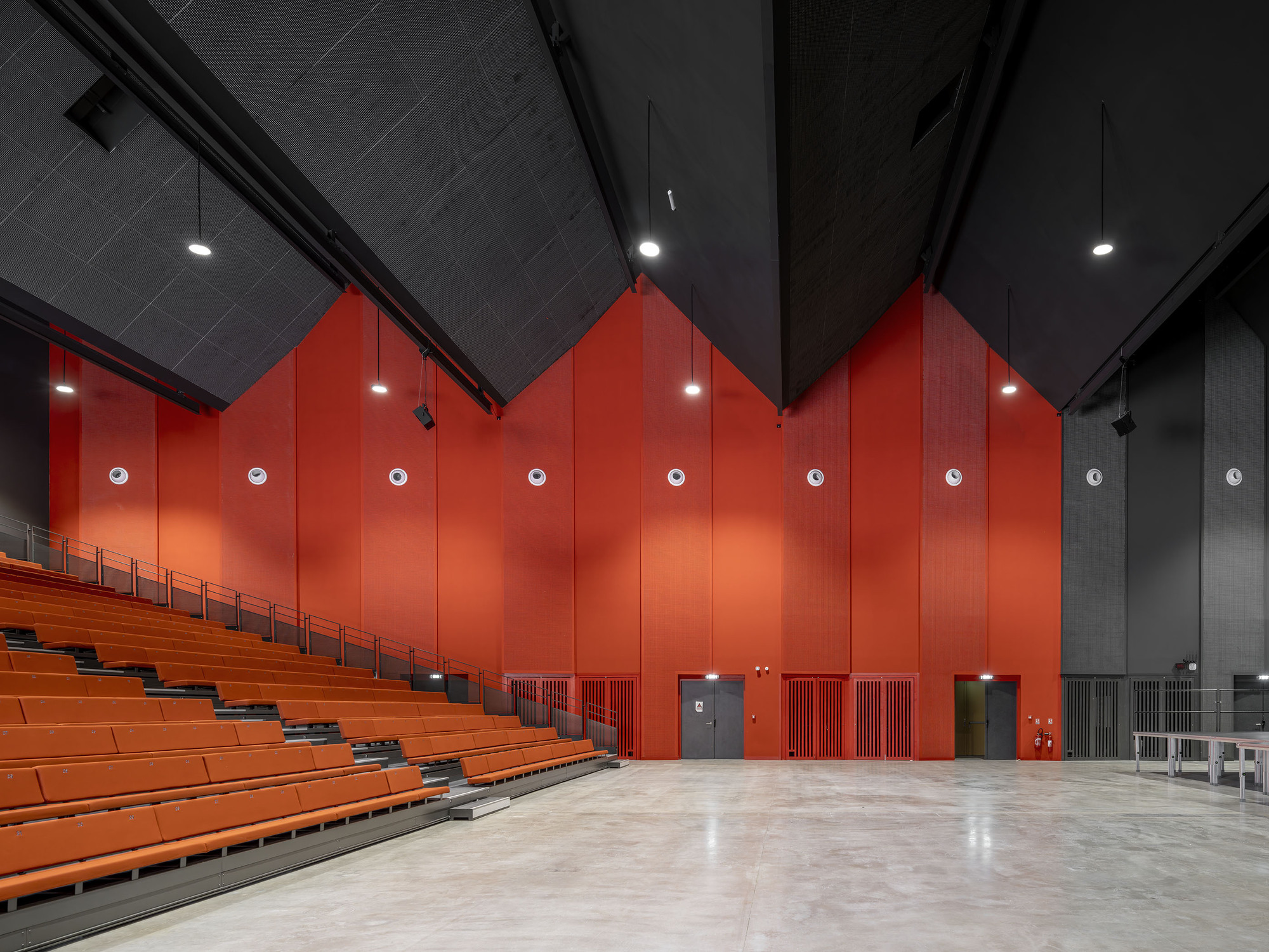 Performance hall with orange tiered seating, a red serrated rear wall and black angled ceiling planes above a polished concrete floor