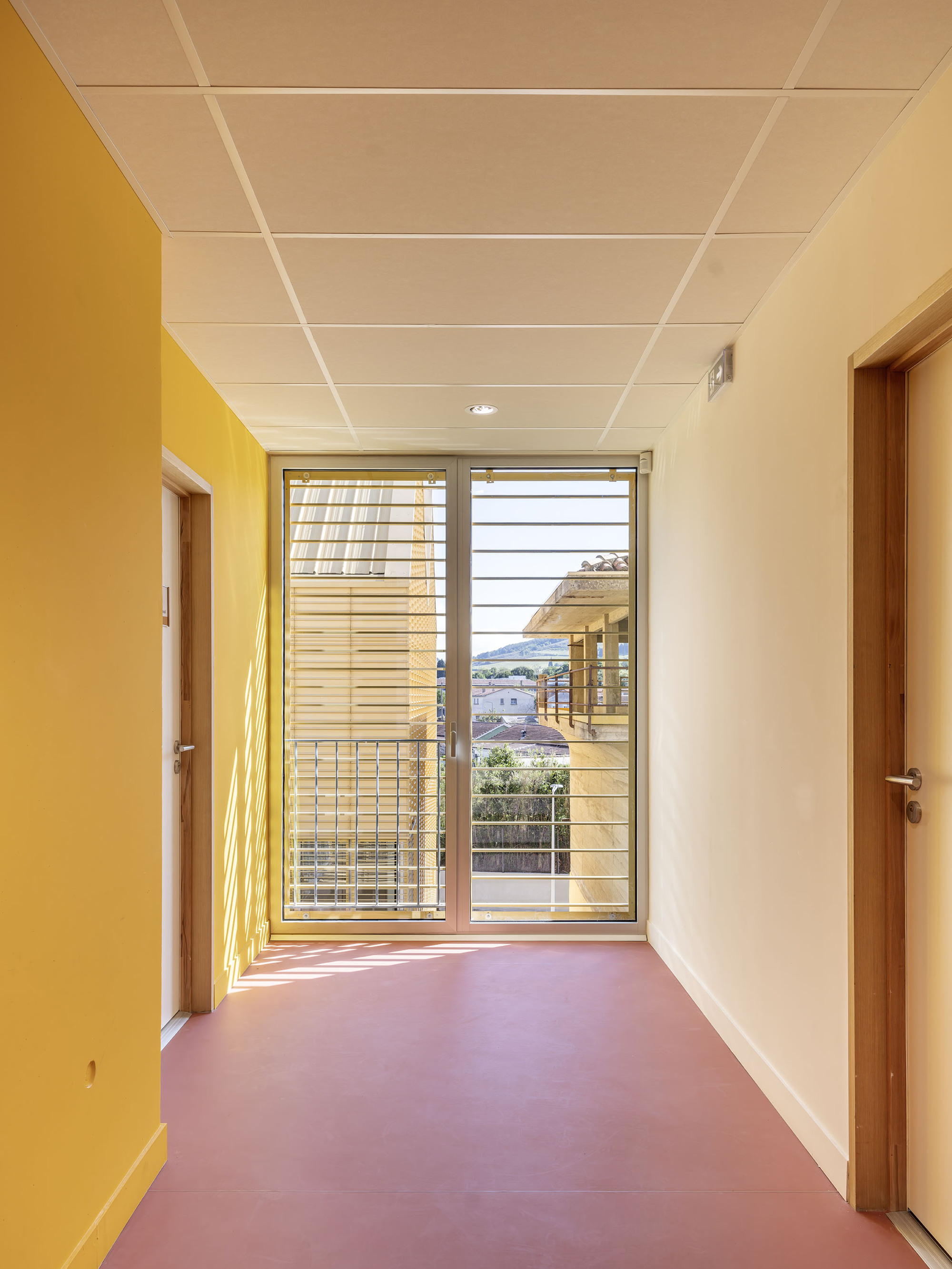 Upper-floor corridor with yellow walls, red floor and a glazed door with horizontal slats, sunlight raking across the floor