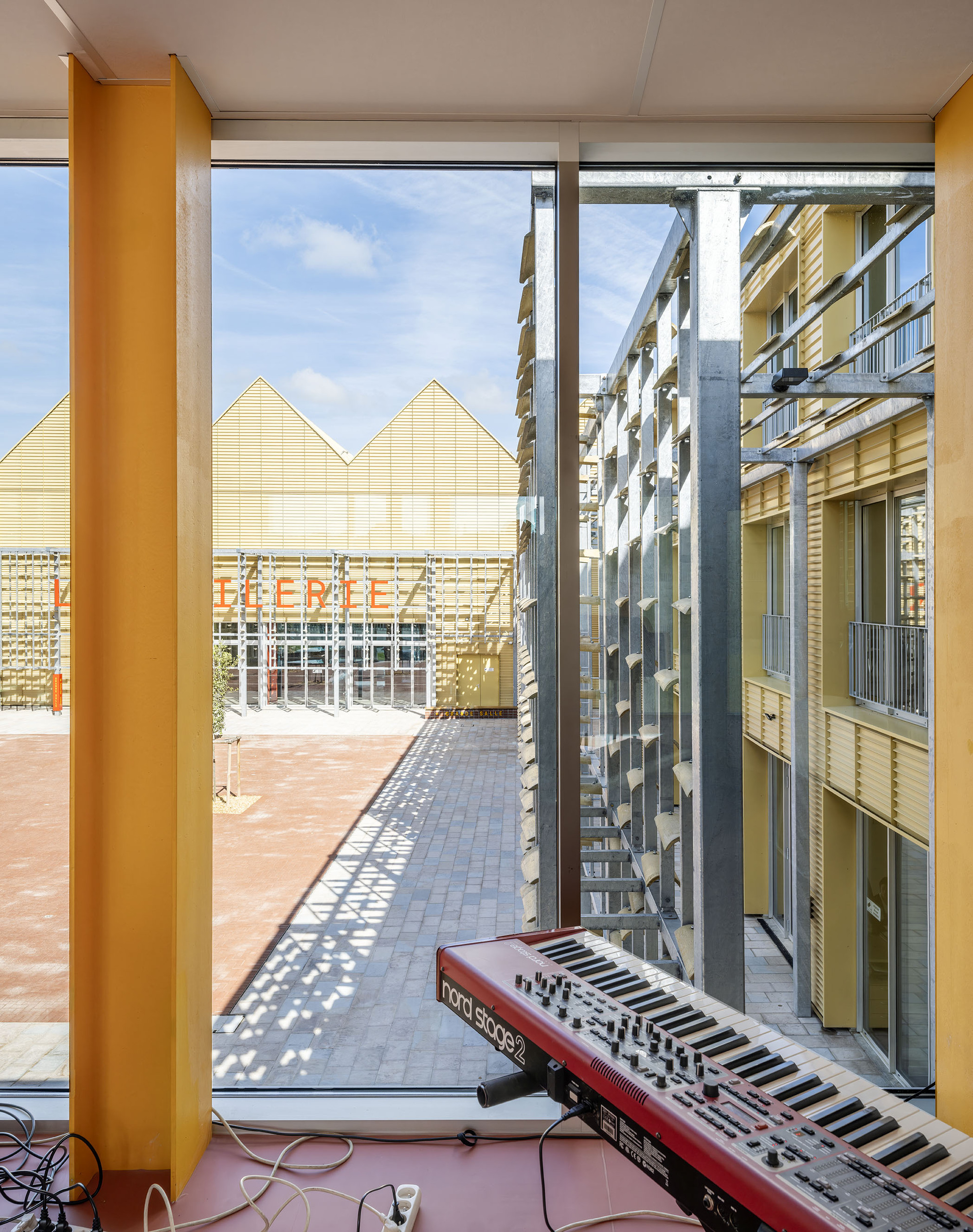View from a music room with a red keyboard toward a sunlit brick courtyard and sawtooth-roofed yellow hall