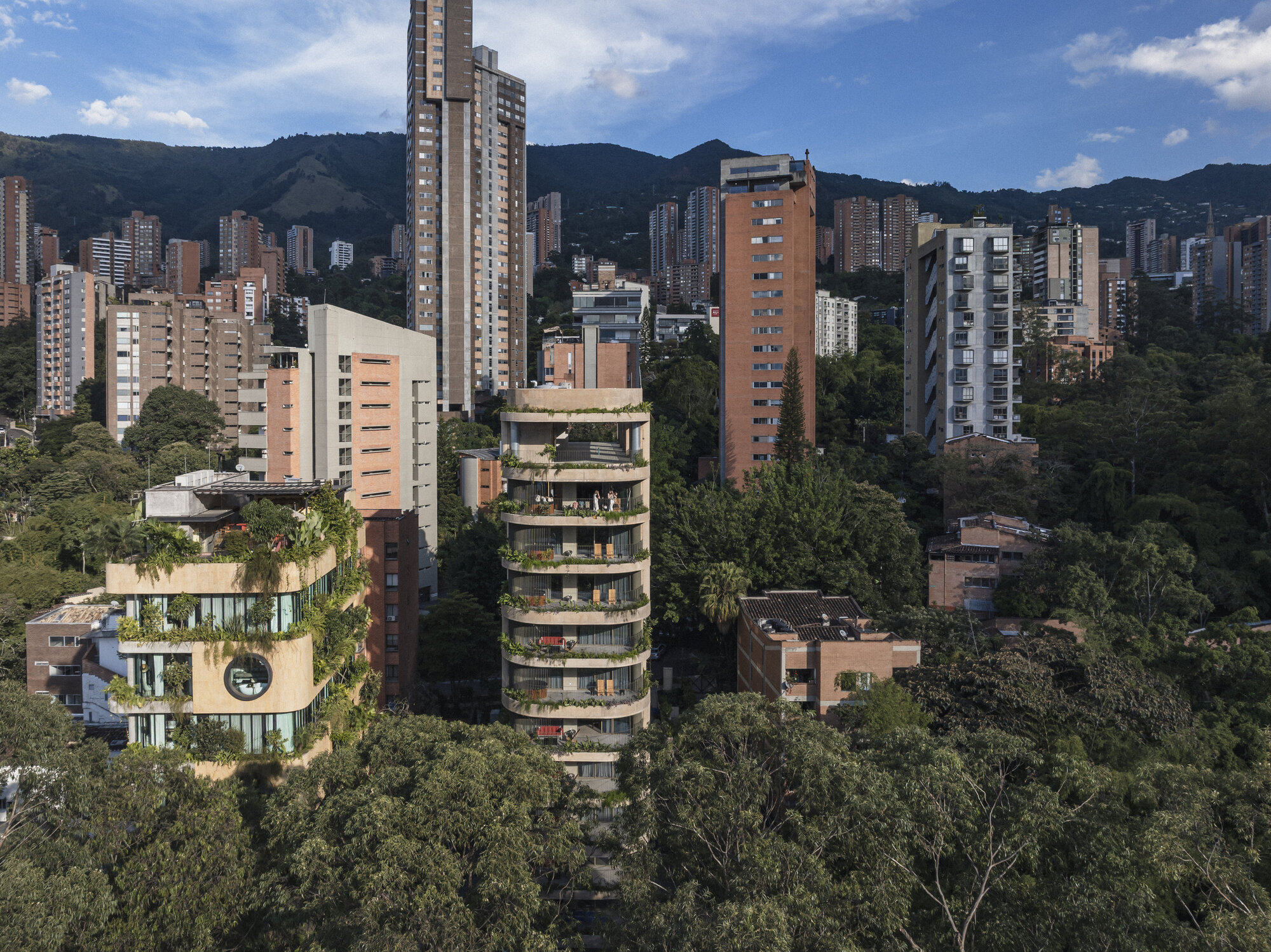 Aerial view of a tower with planted curved balconies among treetops and high-rises, with green hills behind under scattered clouds