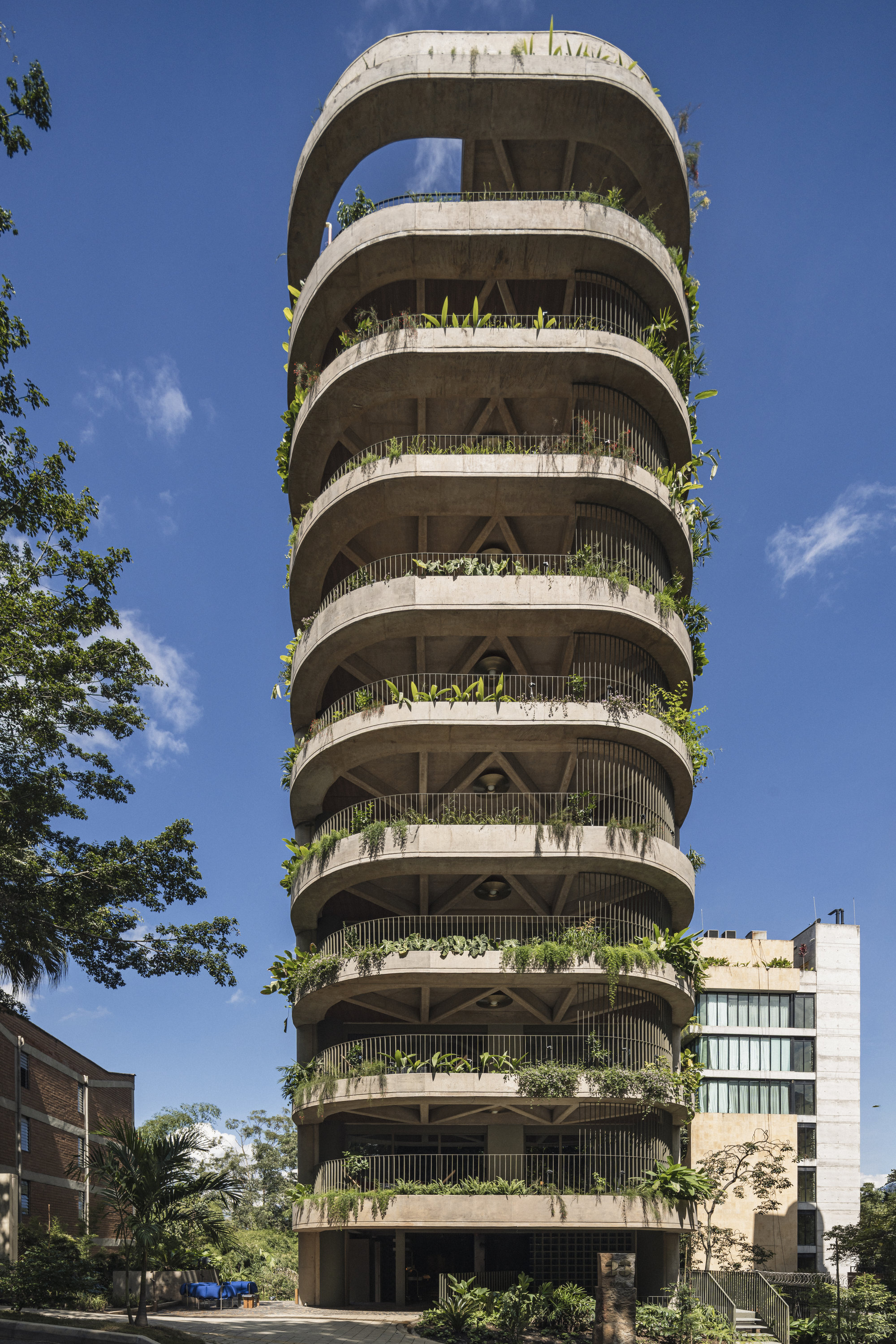 Street-level view of the rounded concrete tower with stacked balconies trailing plants against a deep blue sky