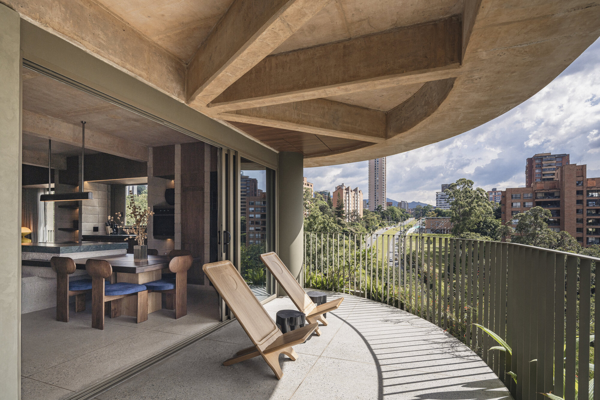 Curved balcony with two wooden folding chairs beside a dining area, exposed concrete beams overhead and city towers in the distance