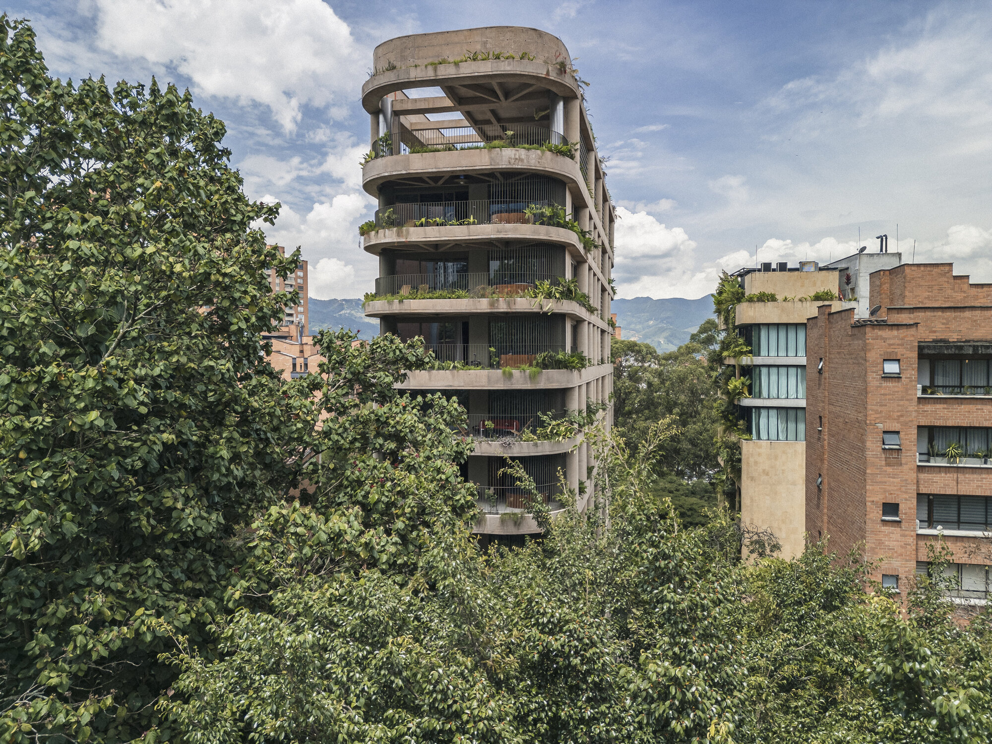 Tower with planted curved balconies rising above a canopy of leafy trees, a brick building beside it under a cloudy sky