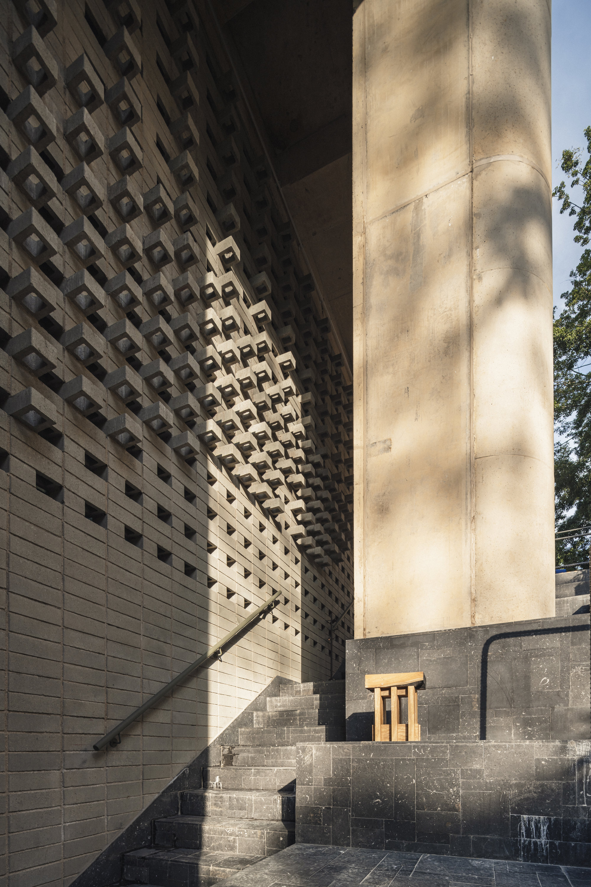 Perforated brick wall beside a rounded concrete column and dark stone steps, with leaf shadows falling across the surface in low sun