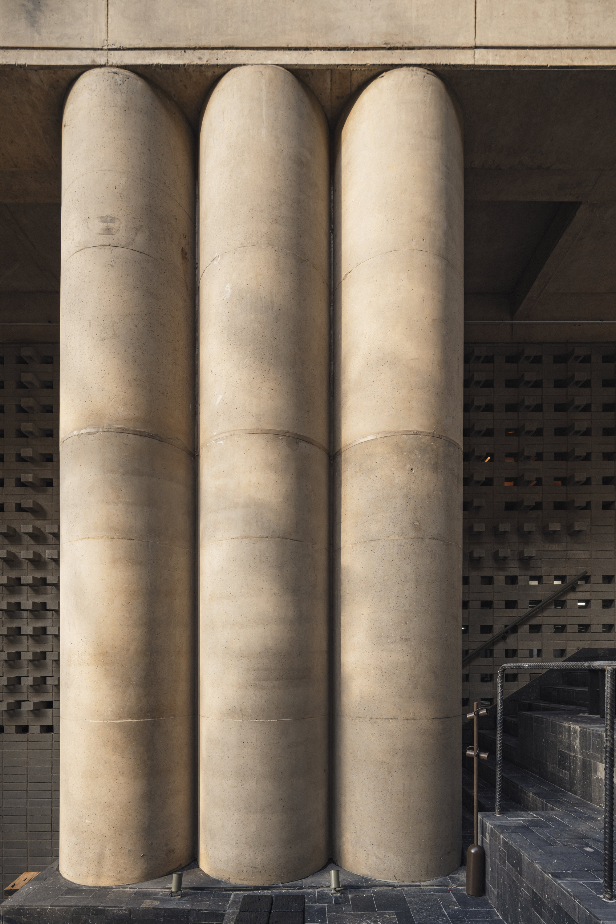 Three rounded concrete columns standing side by side in front of a lattice brick wall and dark tiled steps