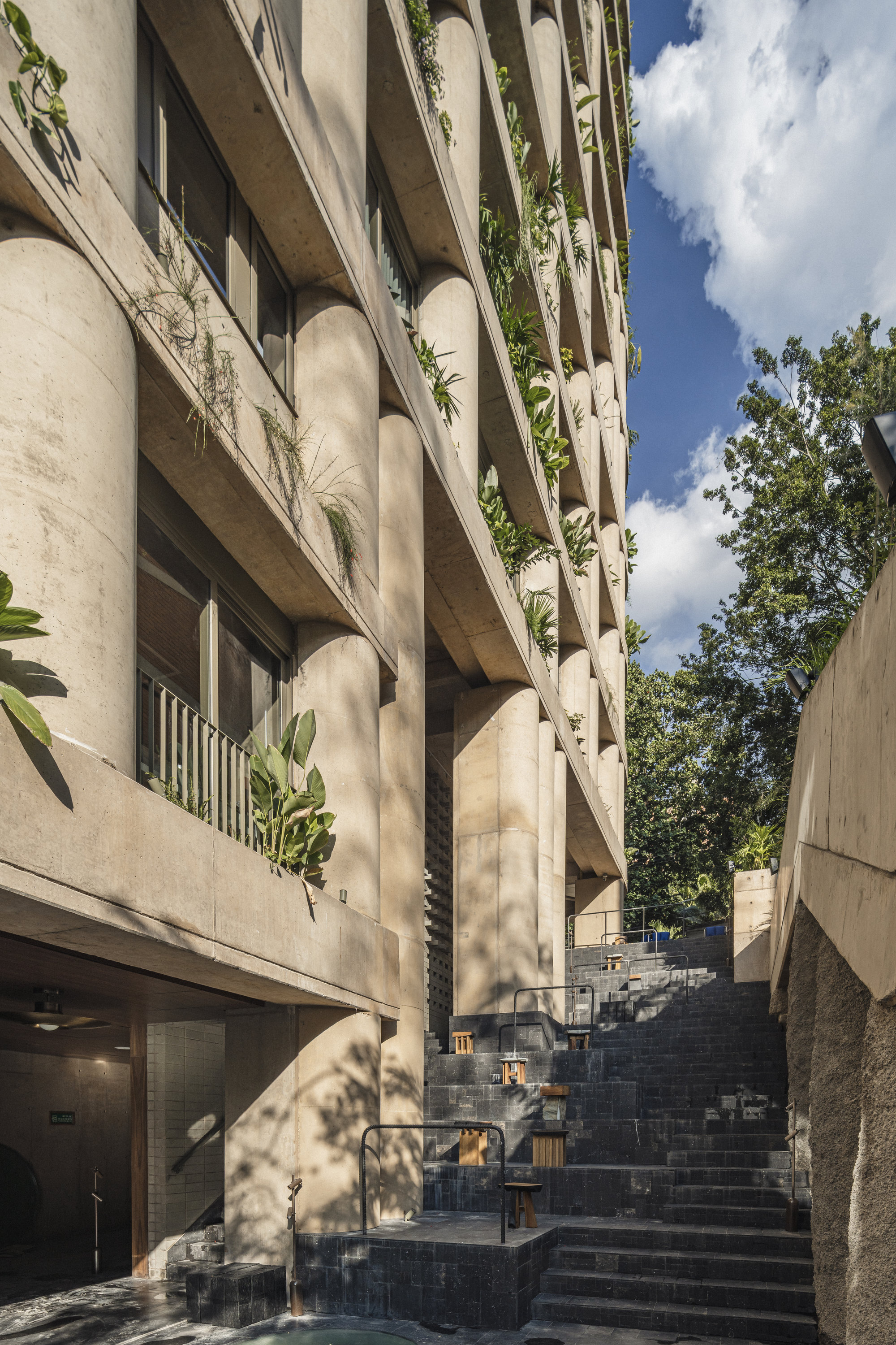 Stepped stone stairway beside a concrete facade of round columns and balconies trailing plants, under a blue sky with clouds