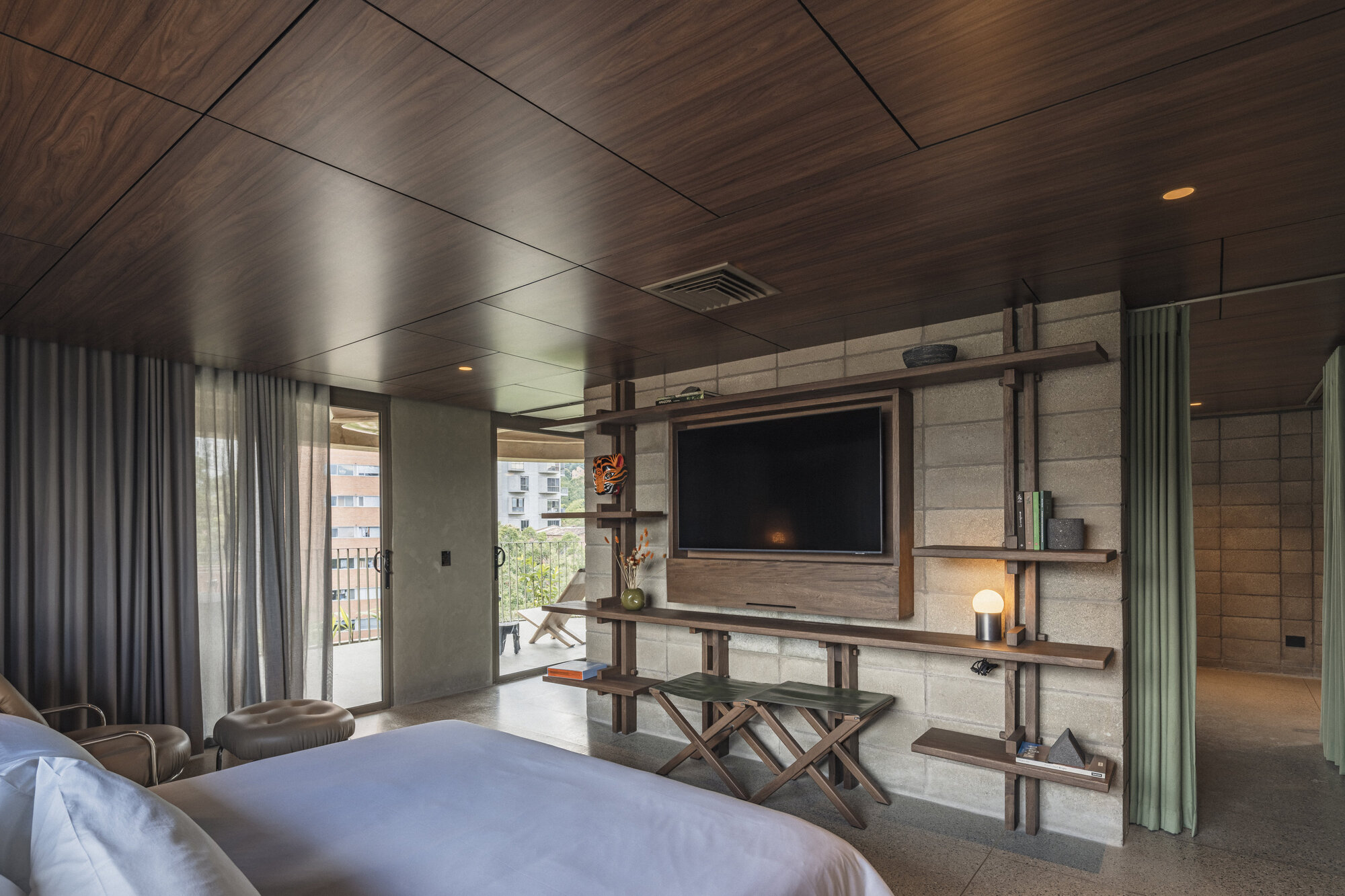Bedroom with walnut ceiling panels, a block wall holding a television and timber shelves, and sheer curtains open to a balcony