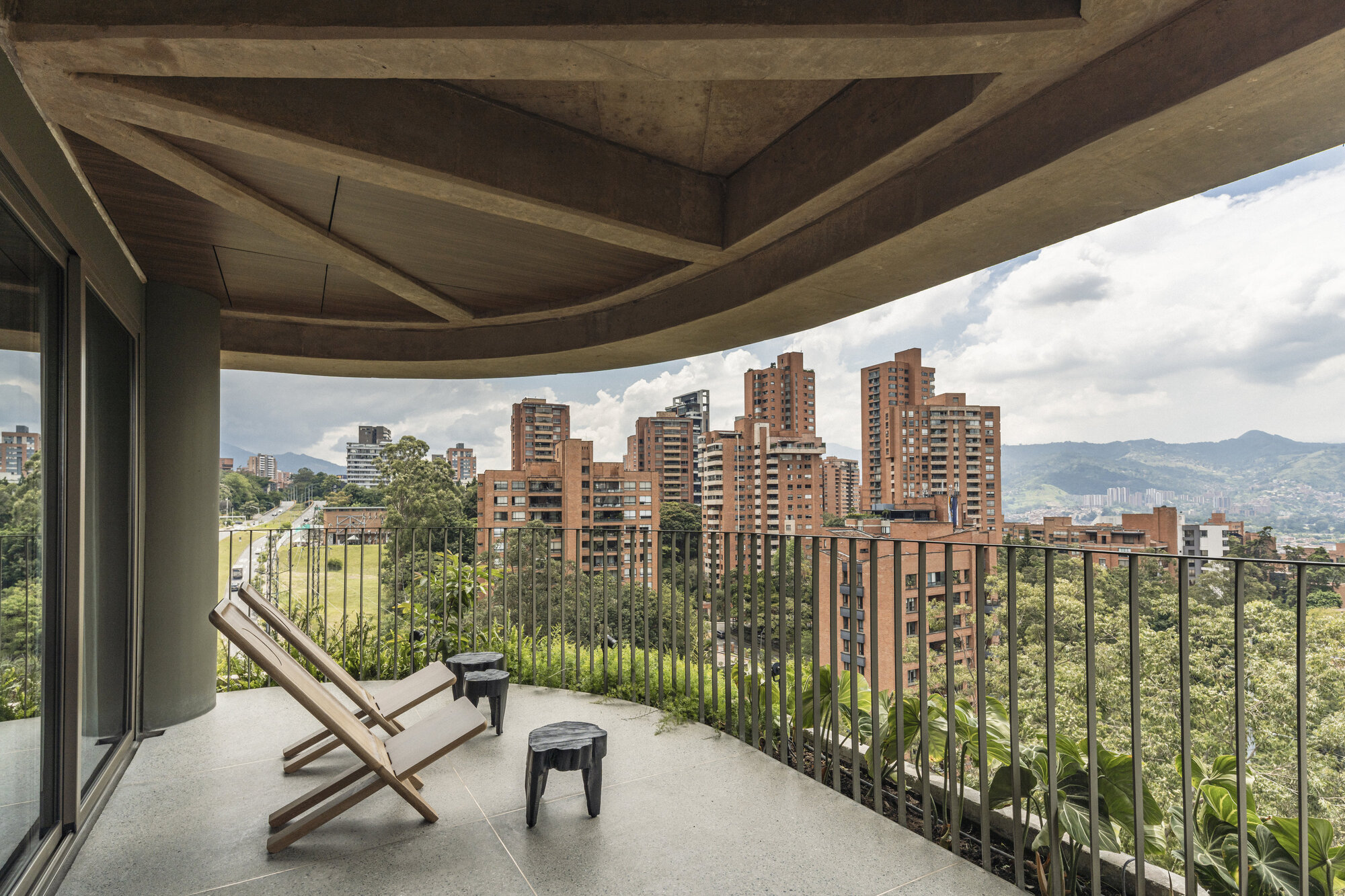 Curved concrete balcony with two timber lounge chairs and black stools, overlooking apartment towers and green hills under cloudy daylight