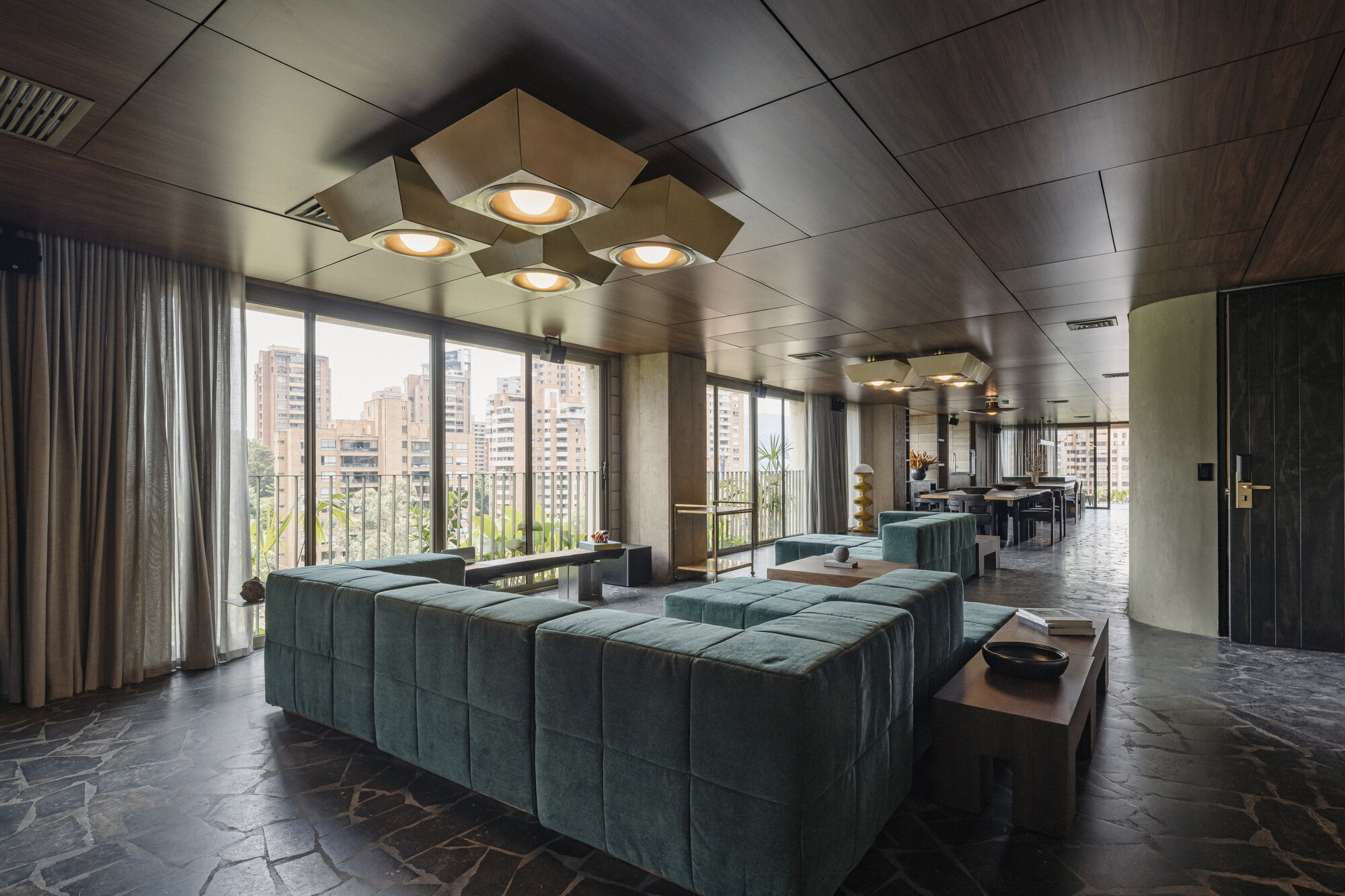 Lounge with green velvet sectional sofas, cluster of faceted ceiling lights and wood-panelled ceiling, floor-to-ceiling windows facing city towers
