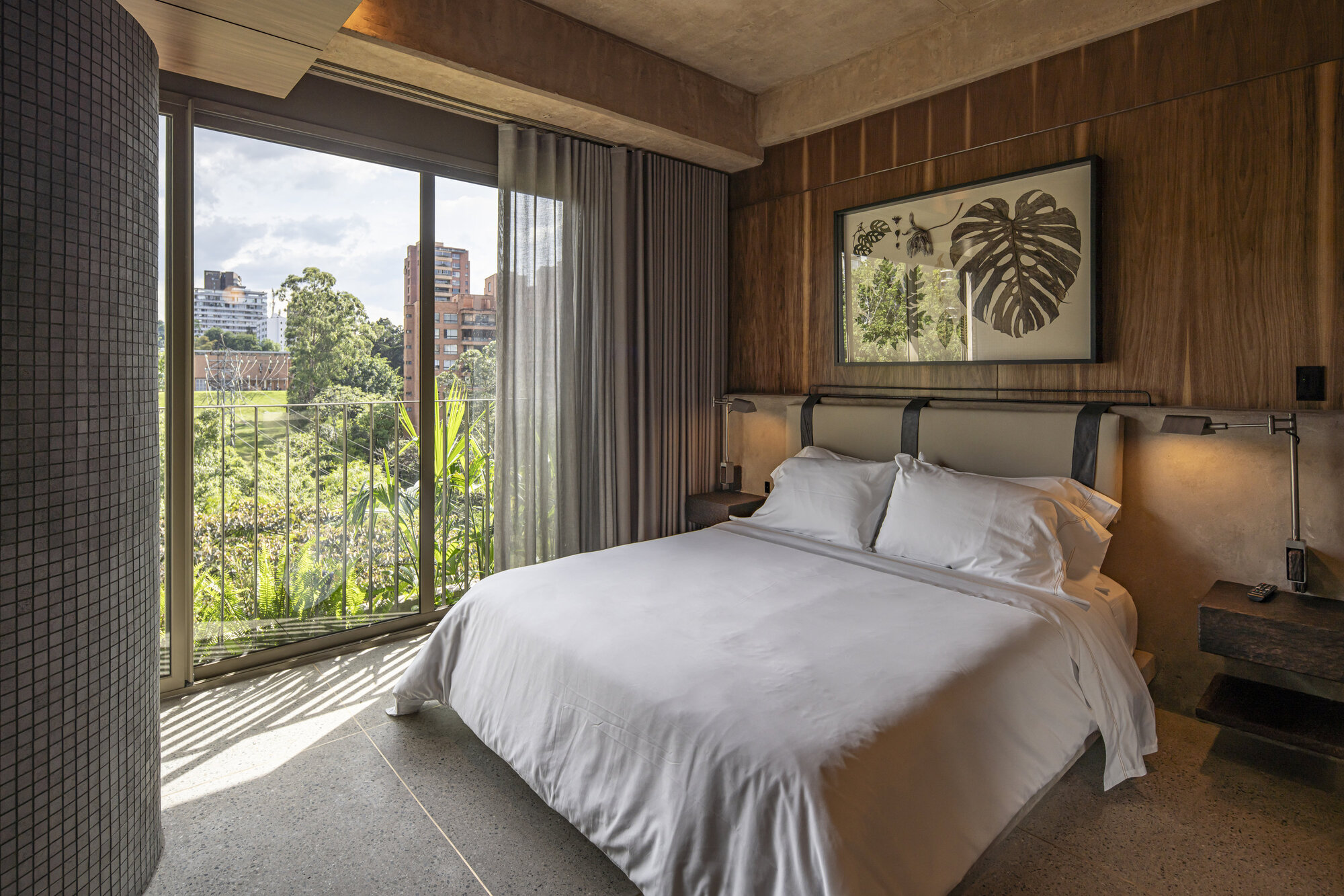 Bedroom with walnut paneled wall, botanical print above the bed and a tiled round column beside a balcony door in afternoon sun