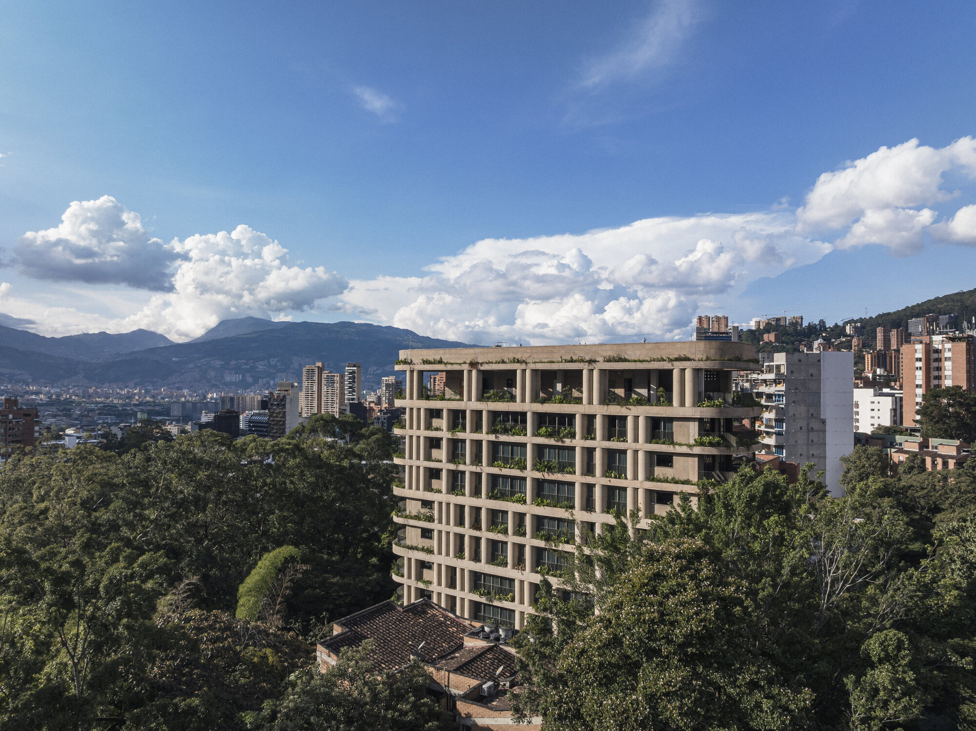 Aerial view of the concrete-framed apartment block with planted terraces, rising above dense tree canopy under towering cumulus clouds