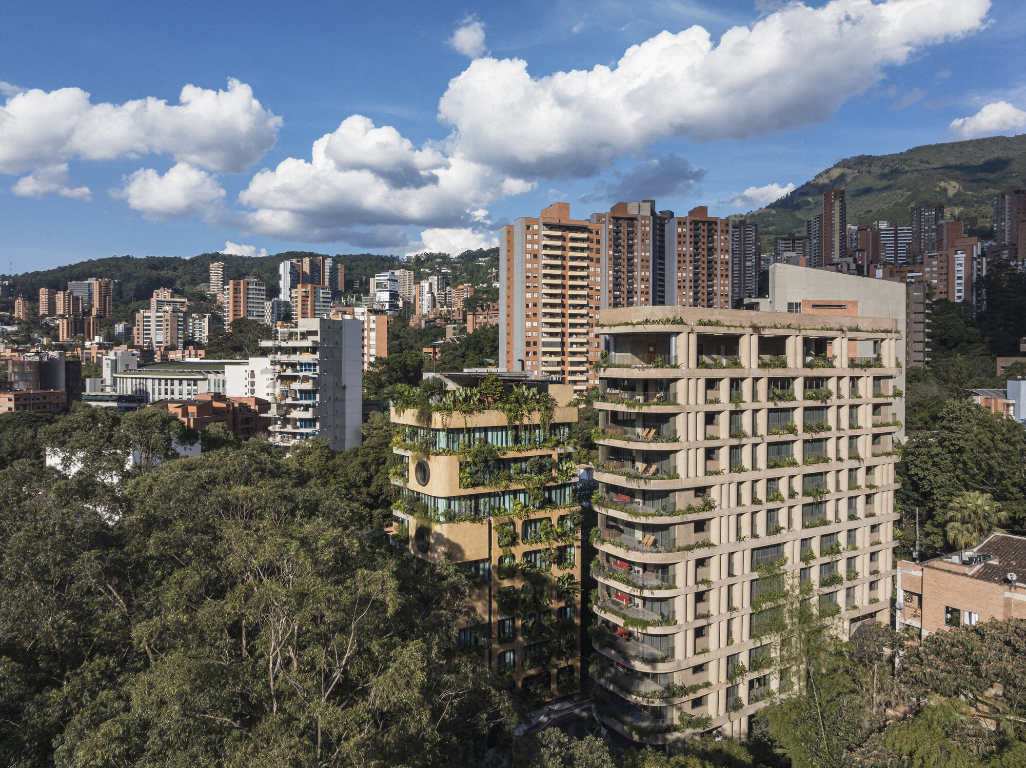 Two stepped towers with planted balconies and curved slab edges, seen above treetops against a blue sky and hillside
