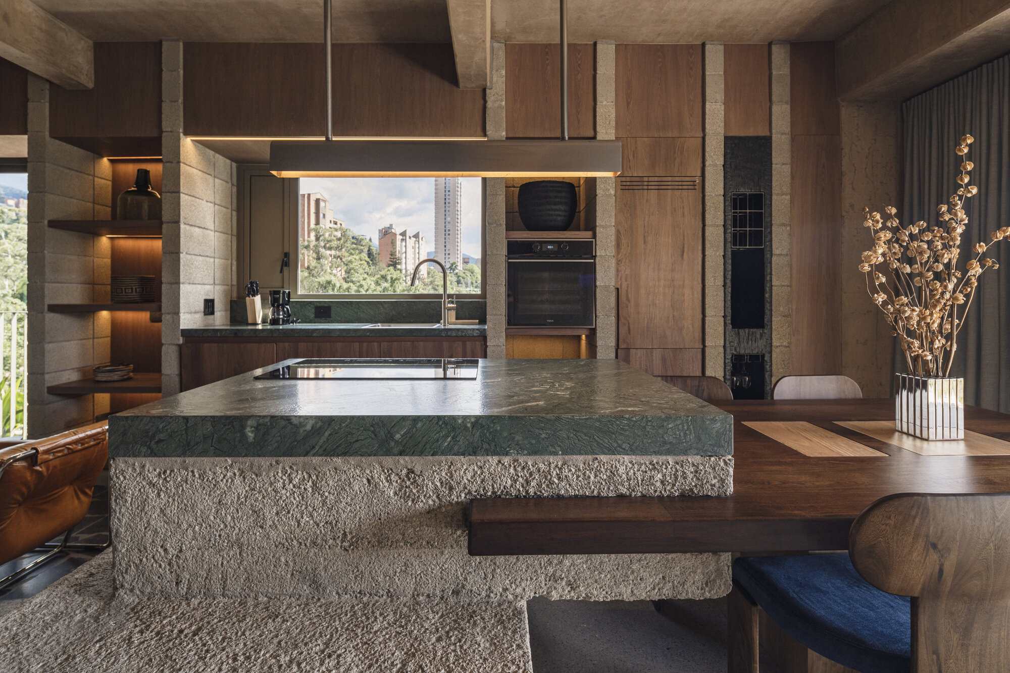 Kitchen island with green stone countertop on a rough-hewn stone base, walnut cabinetry behind and a dried-flower vase on the table