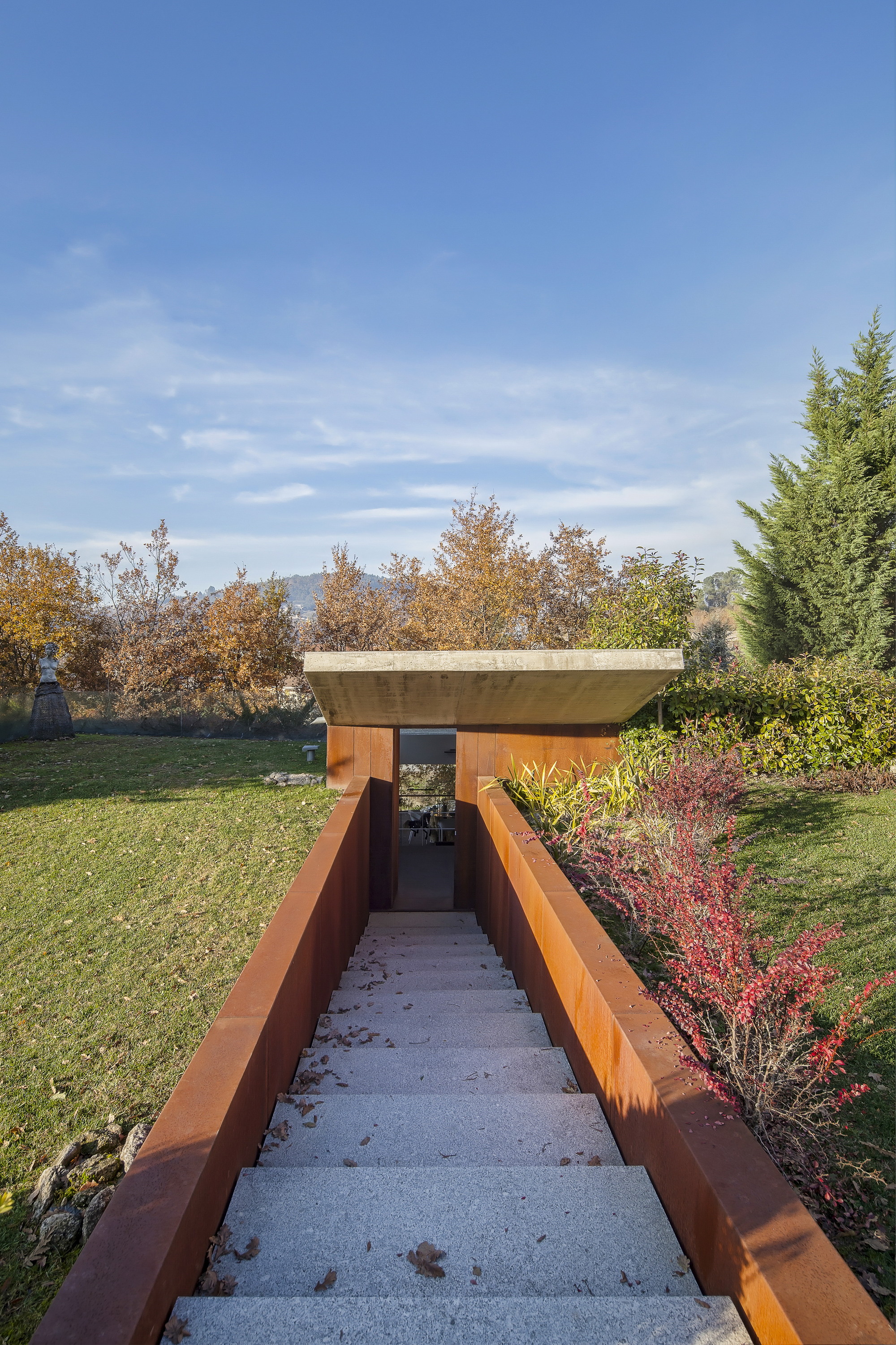 Granite steps between weathering steel walls leading down to a concrete-roofed entrance, with red shrubs and a lawn in autumn