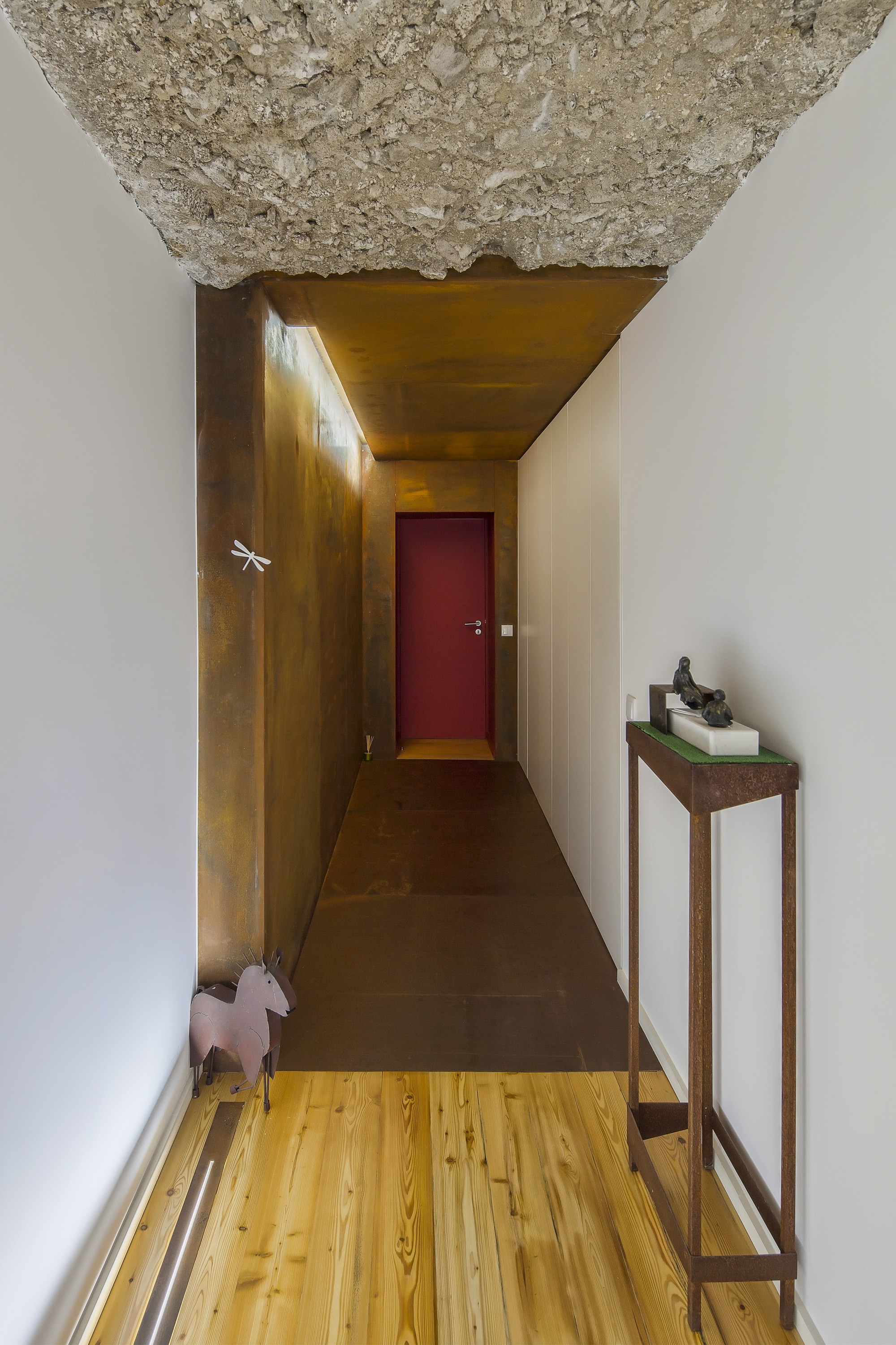 Narrow corridor with a rough concrete ceiling, rusted steel wall and red door at the end, over a pine floor
