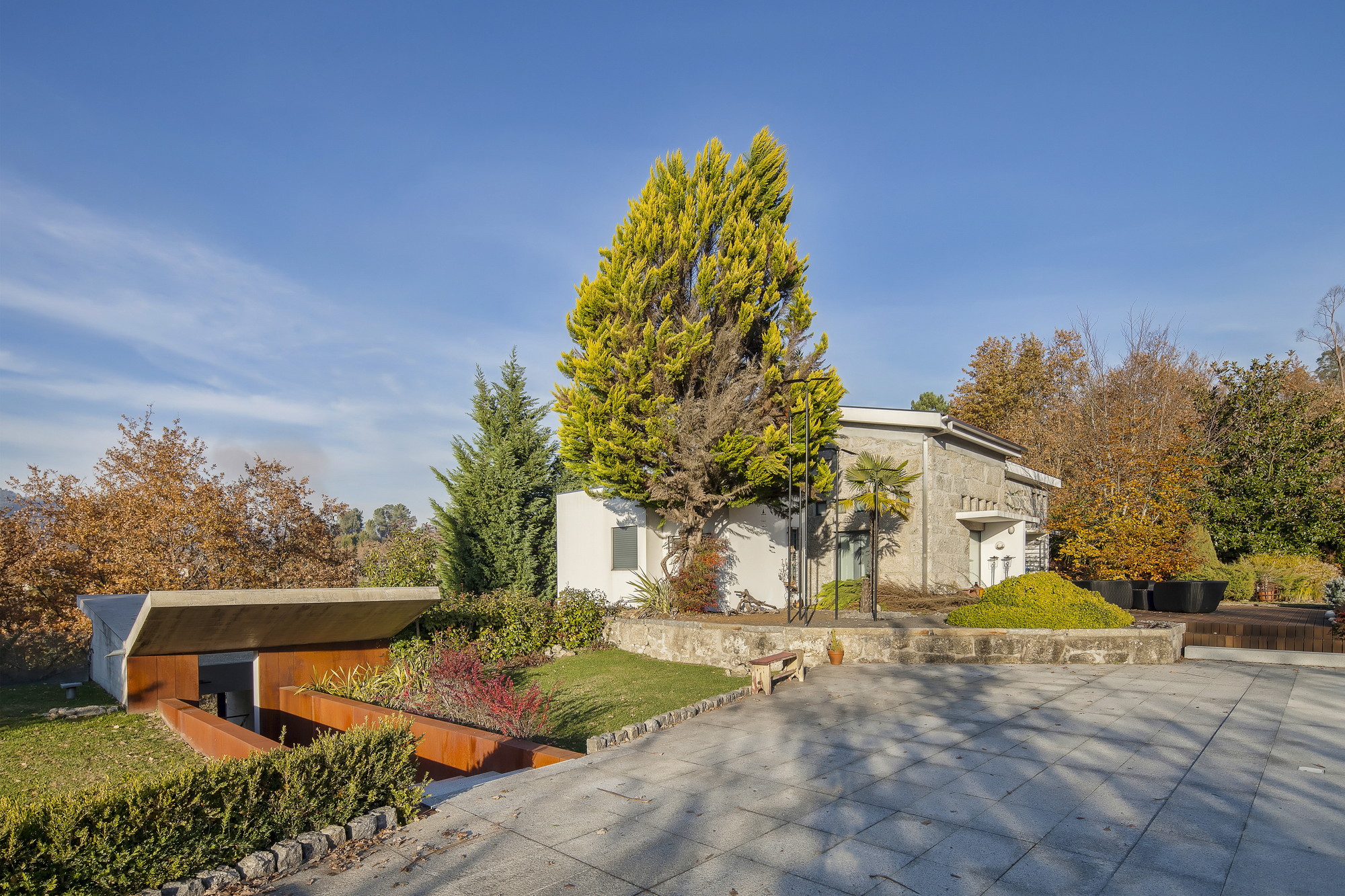 Granite paved terrace and weathering steel planter walls beside a low sloped roof, with a yellow-green conifer in autumn light