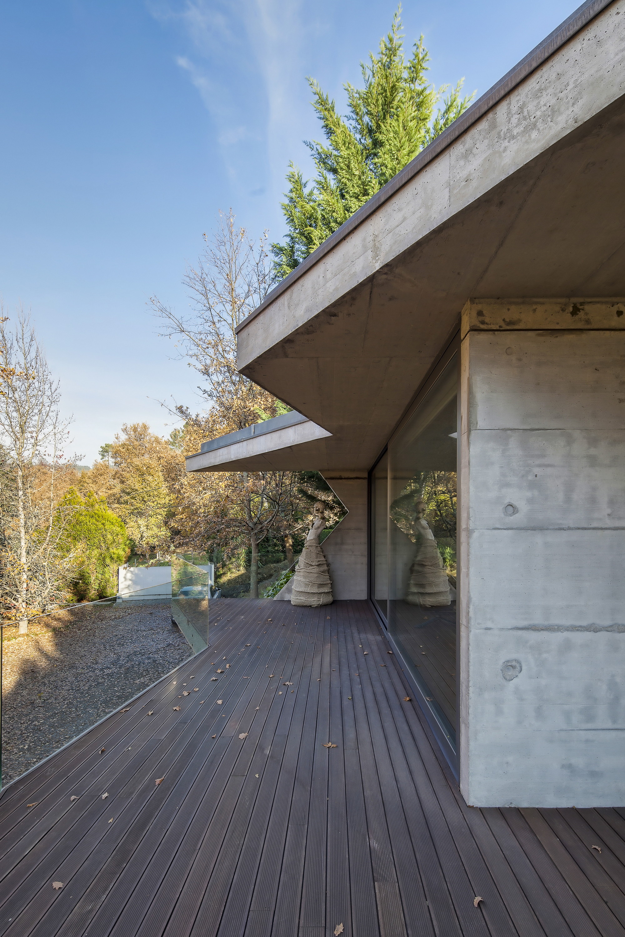 Timber deck running along a board-marked concrete wall and overhanging slab, with glazing reflecting trees under a clear blue sky
