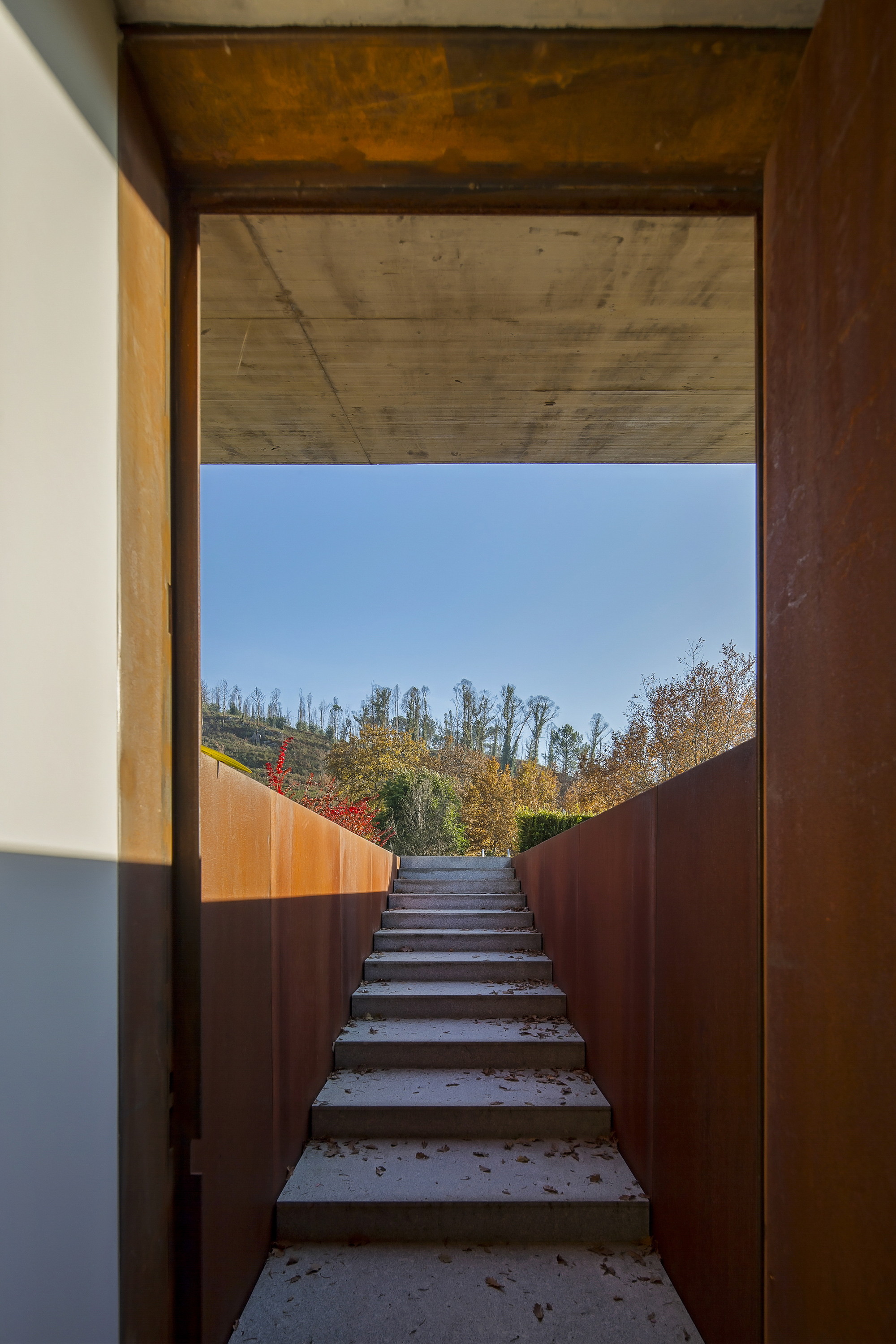 Concrete stair rising between weathering steel walls toward a framed view of autumn hillside, with fallen leaves on the steps