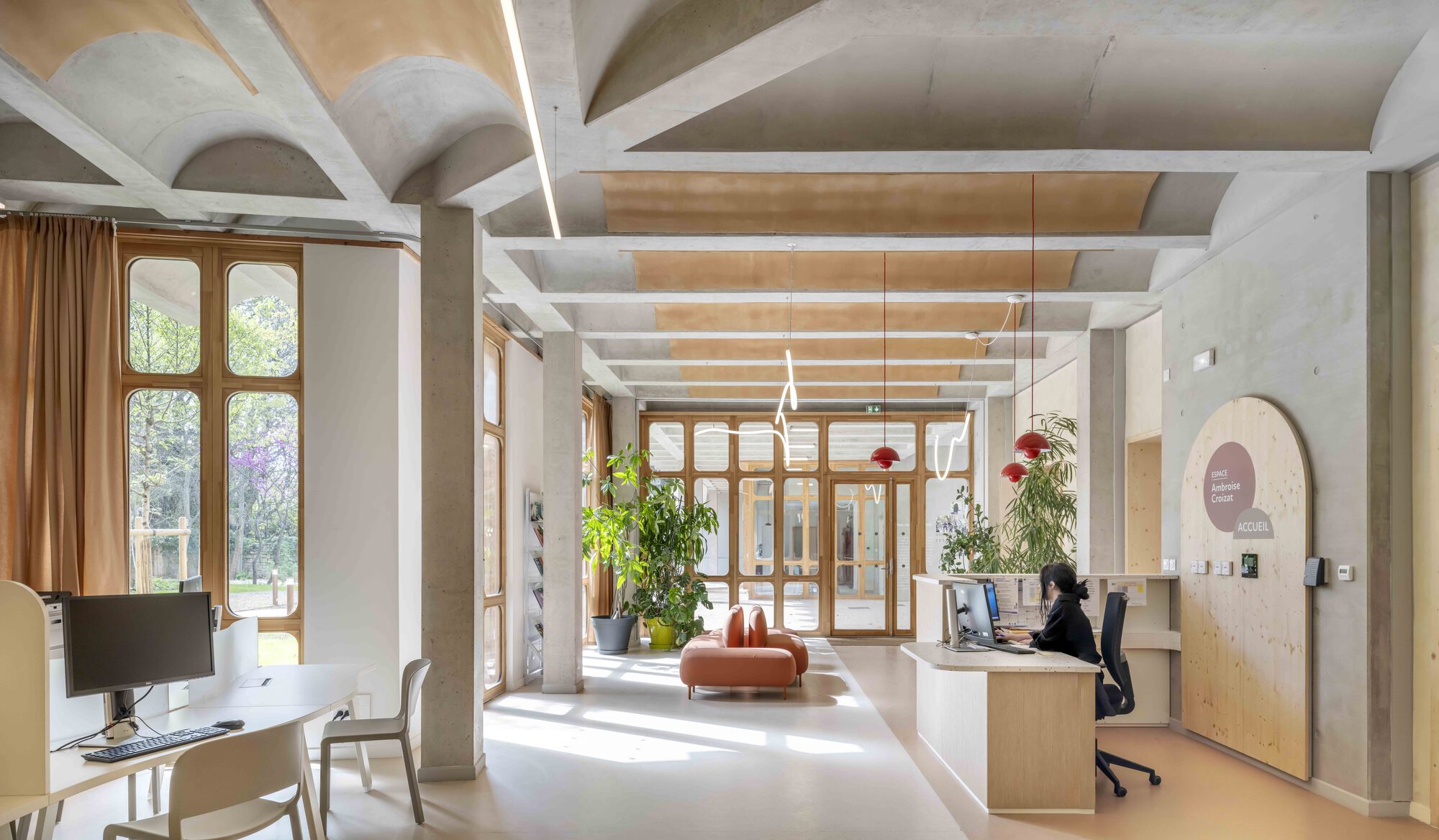 Reception hall with concrete vaulted ceiling, timber-framed windows and a curved wooden door, sunlight falling across the floor as a staff member works at the d
