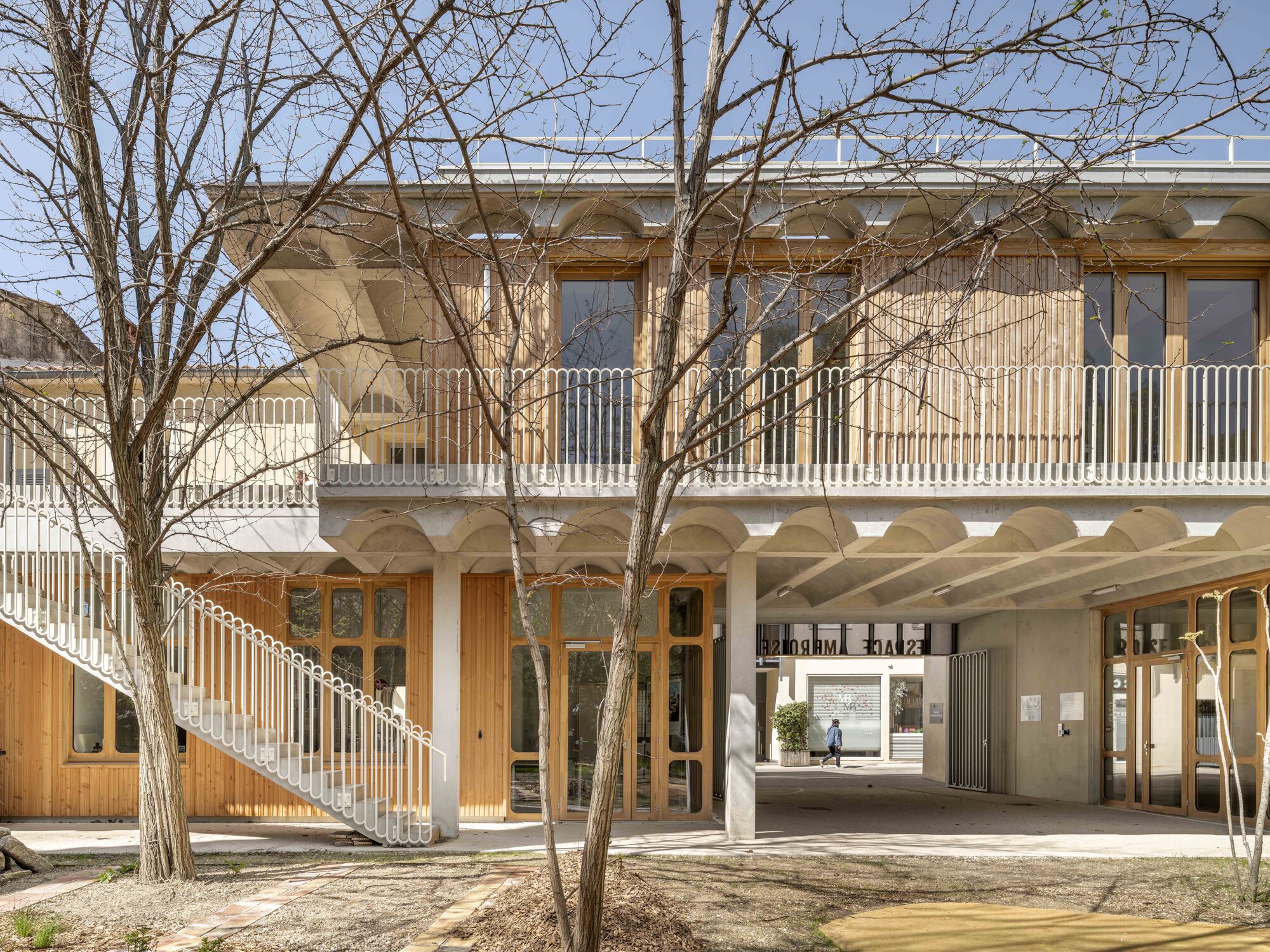 Two-storey courtyard facade with timber cladding, white balustrade and exterior stair, seen through bare branches under a clear blue sky