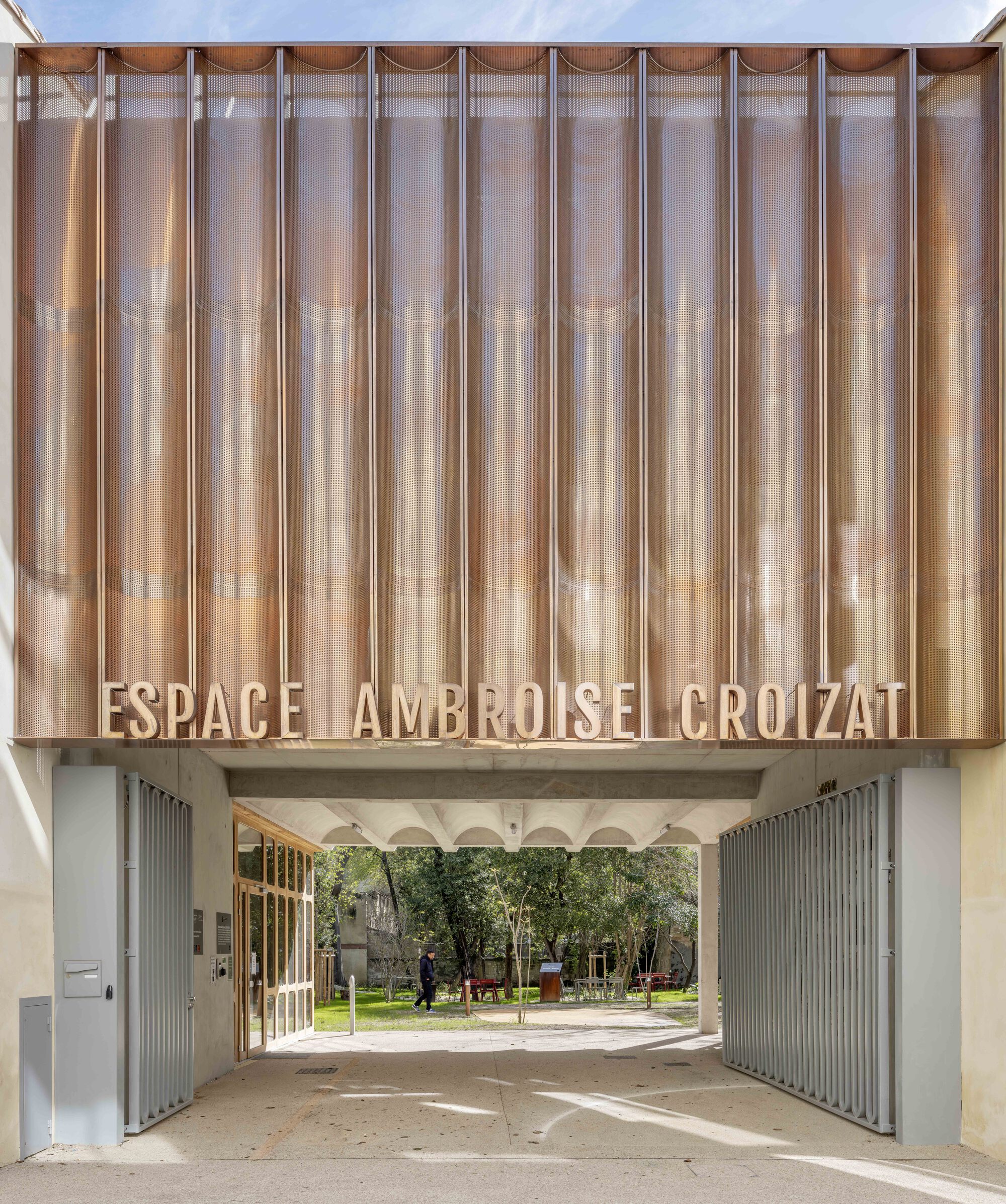 Entrance gateway beneath a copper-toned fluted screen carrying the building name, with a pedestrian walking in the garden beyond