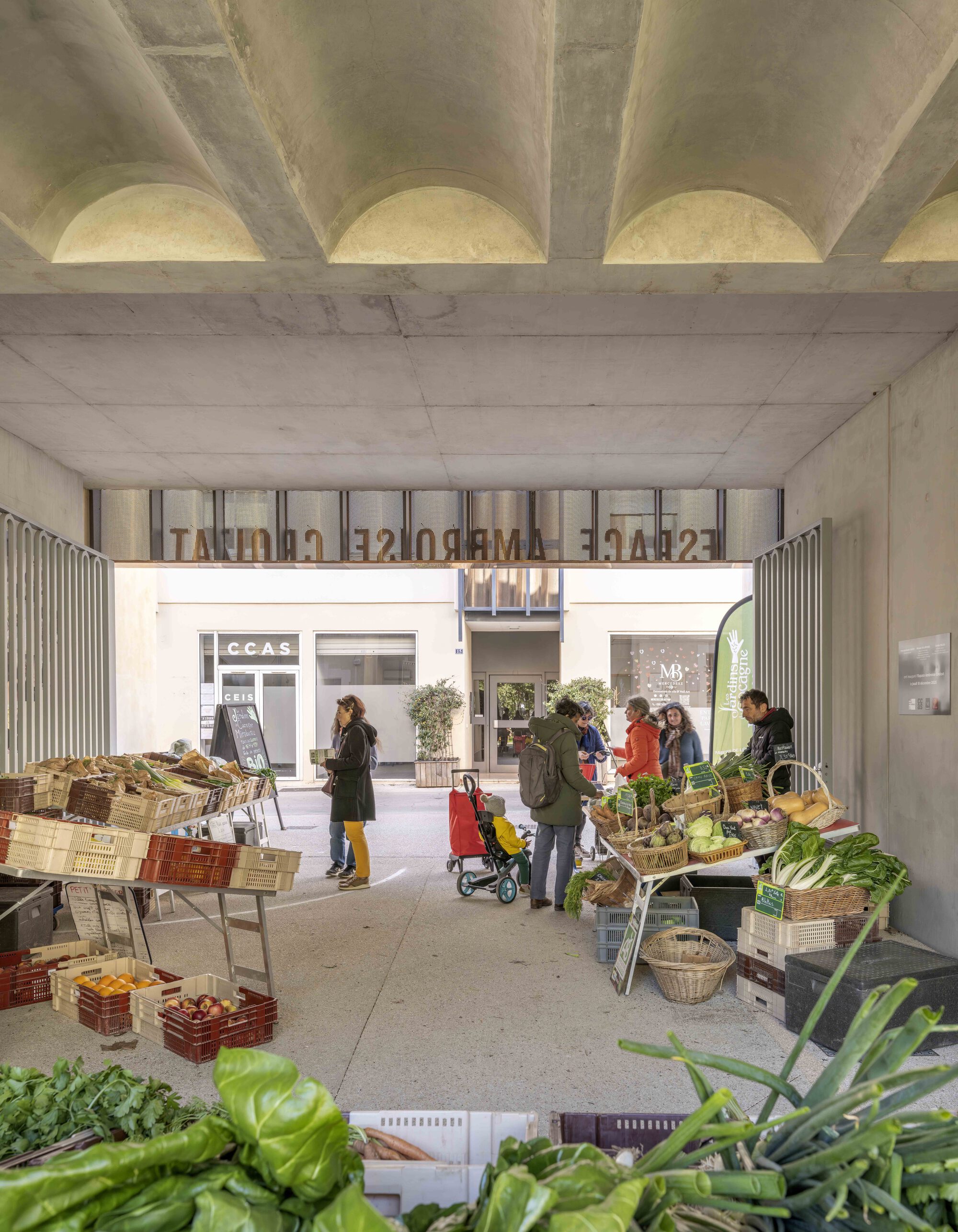 Market stalls of crated vegetables and leafy greens under the concrete vaulted passage, shoppers gathered in the soft daylight