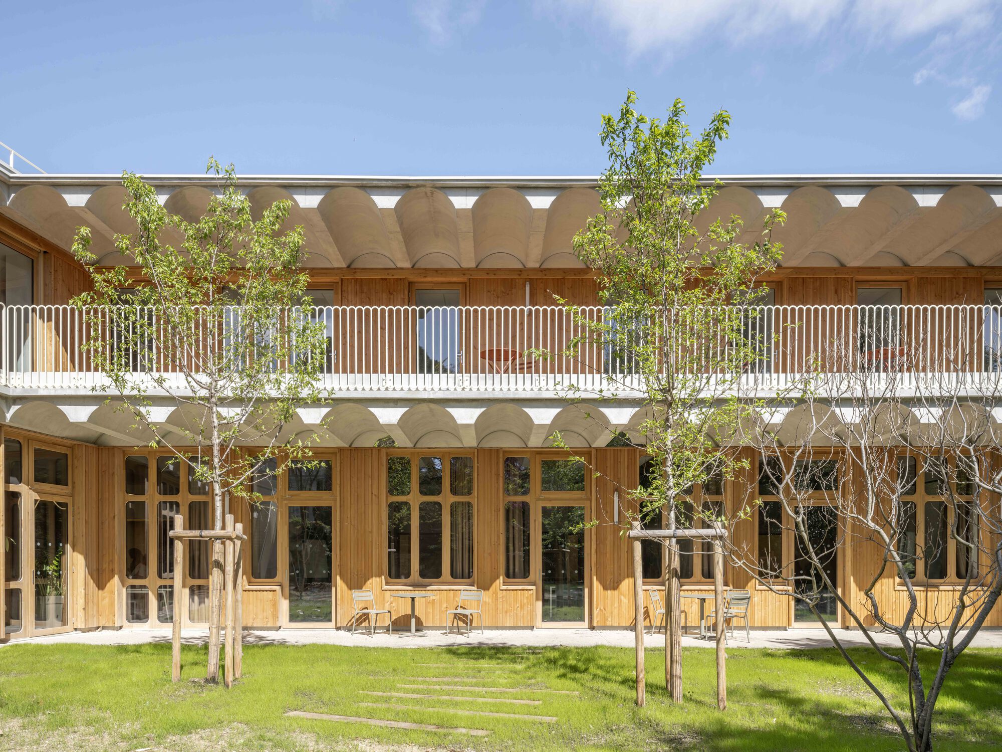 Courtyard elevation with timber-framed glazing, scalloped concrete soffit and young trees staked in the lawn under a bright spring sky