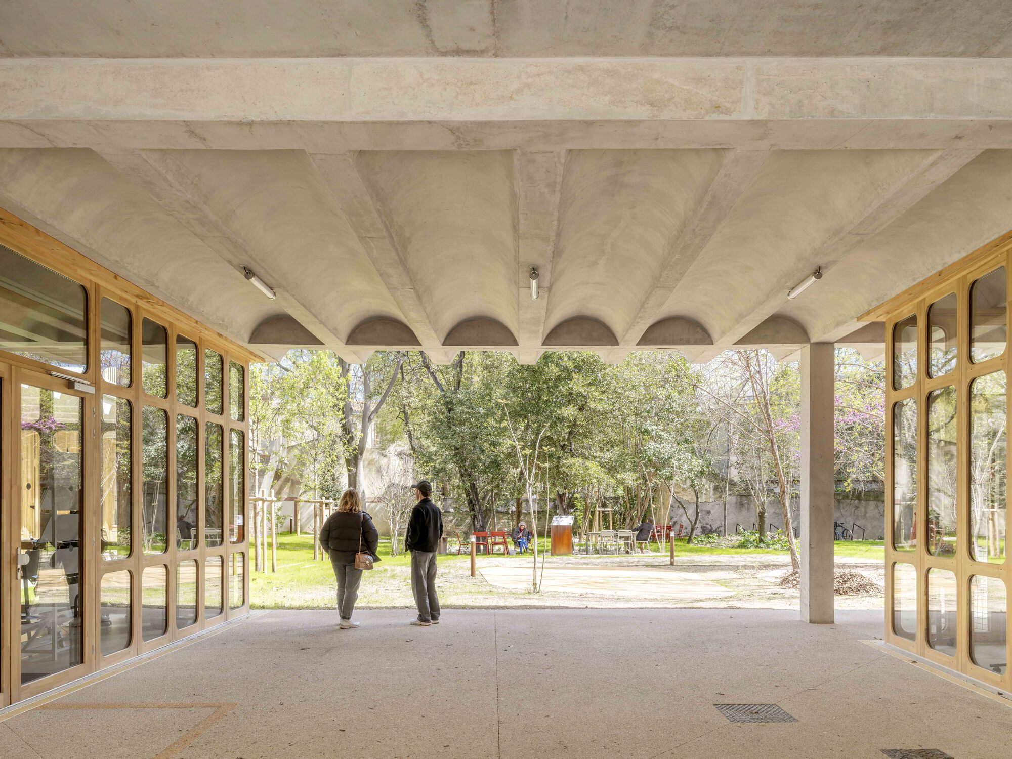 Covered ground-floor walkway under a ribbed concrete vault, with two visitors looking out toward a garden of spring trees