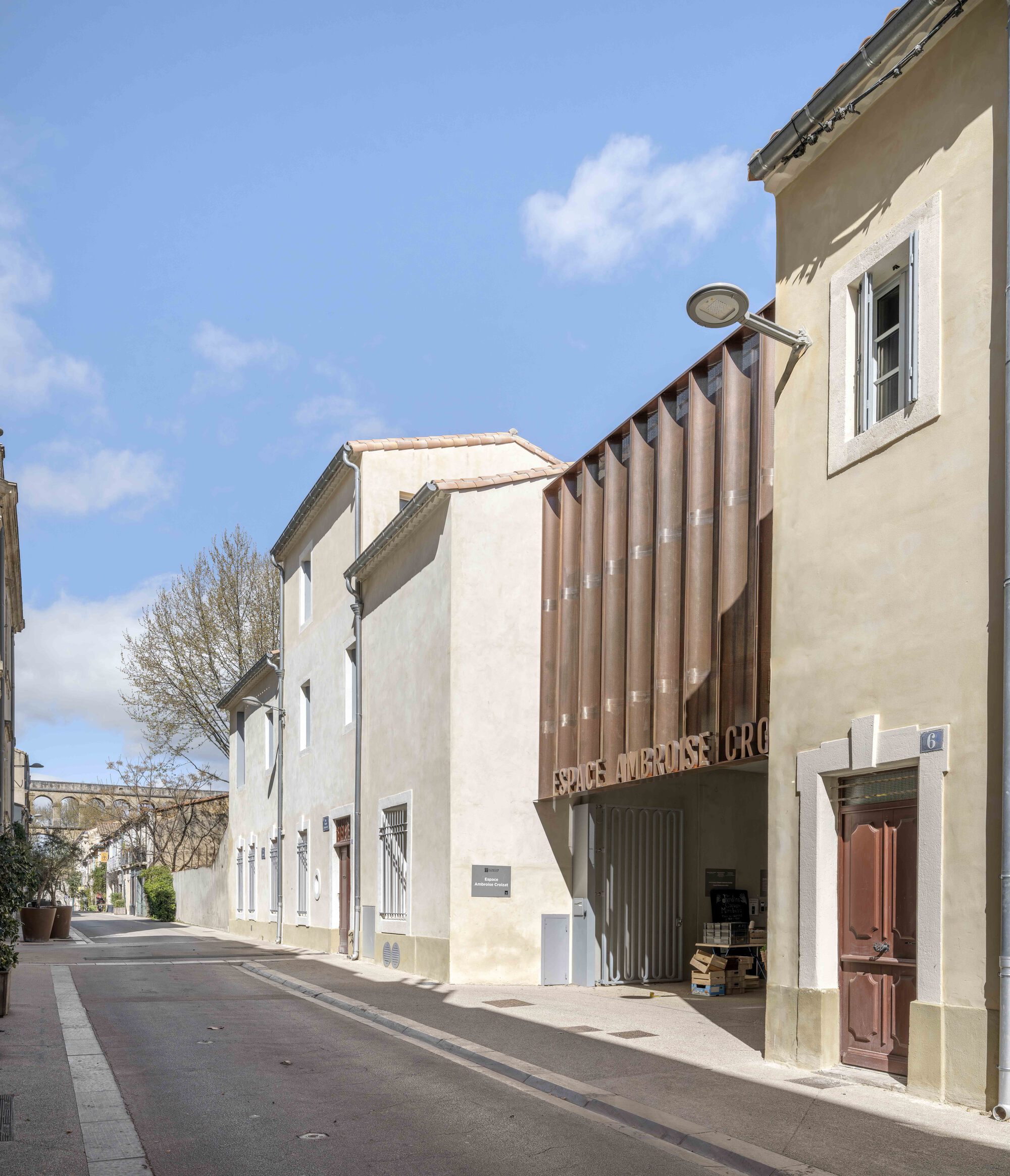 Street view of a copper-toned pleated screen above a gated passage between plastered houses under a blue sky