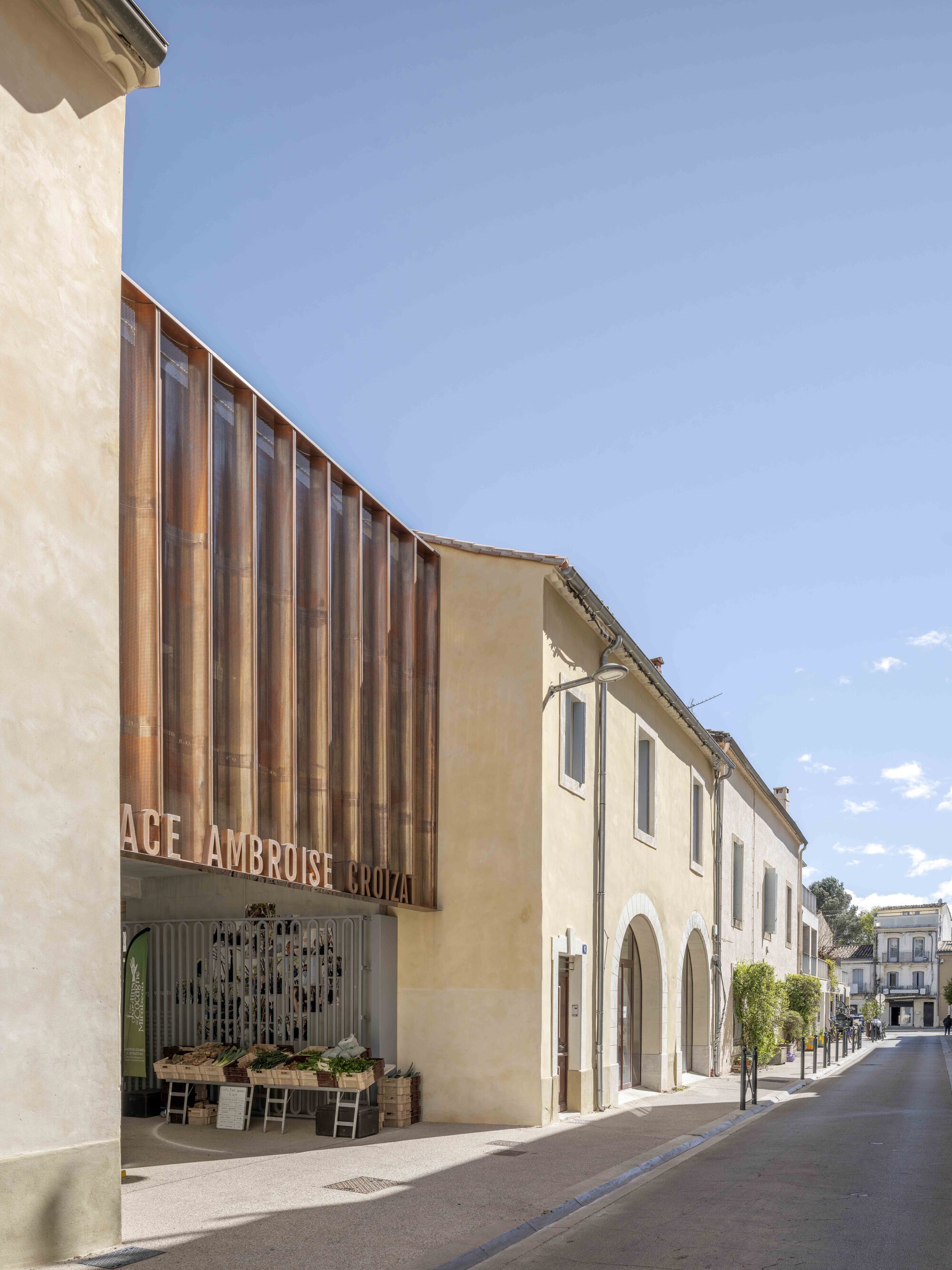 Pleated rust-colored metal facade with lettering above a market stall, beside arched stone houses in afternoon sun