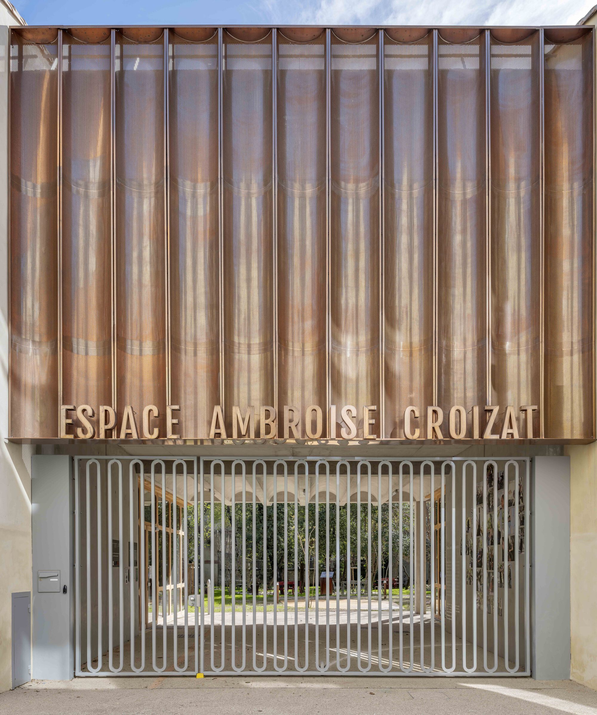 Frontal view of the vertically pleated copper screen over a slatted steel gate opening onto a tree-filled courtyard