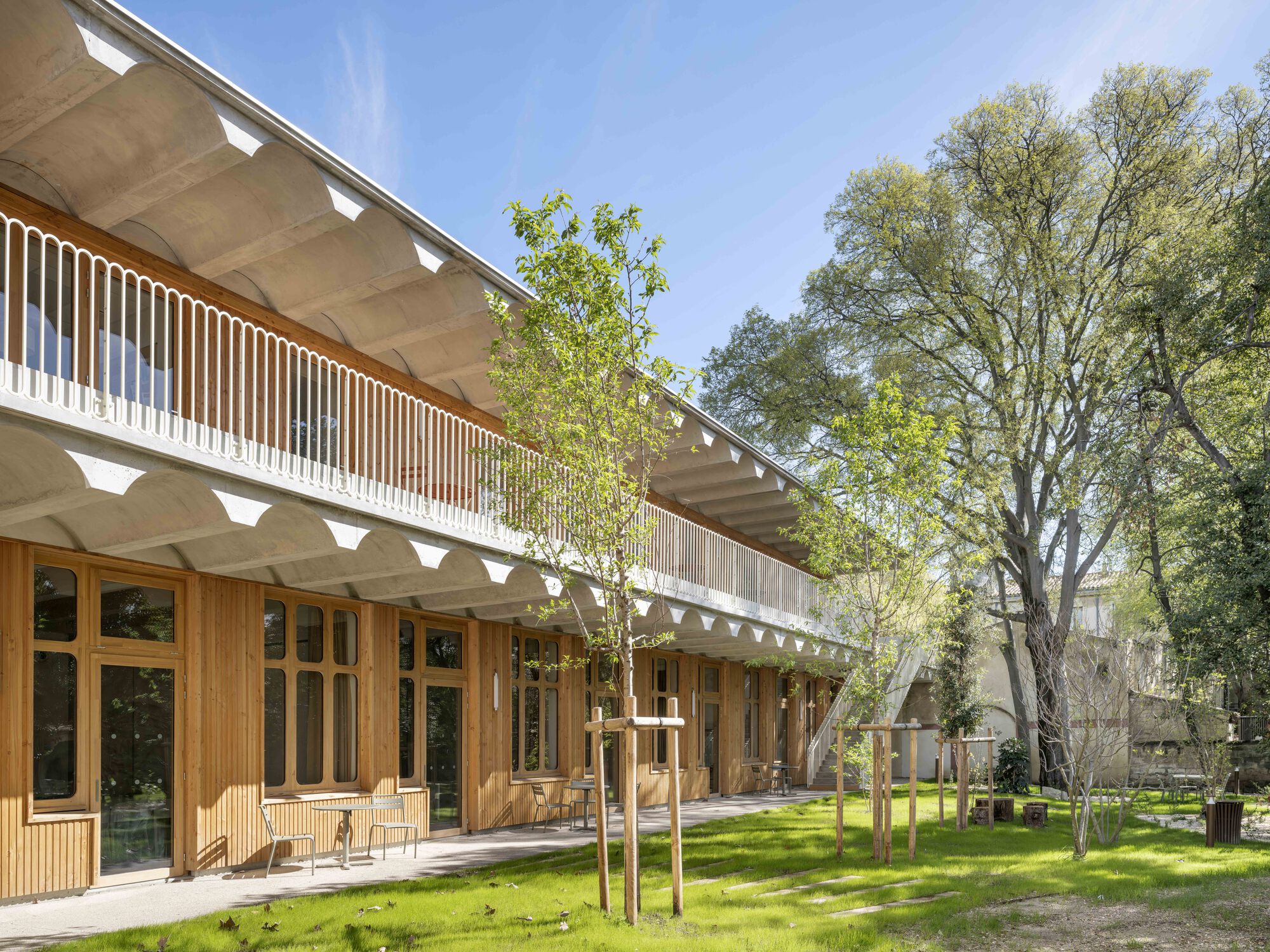 Two-storey timber-clad courtyard wing with a ribbed concrete balcony soffit, young trees and fresh grass in bright sun