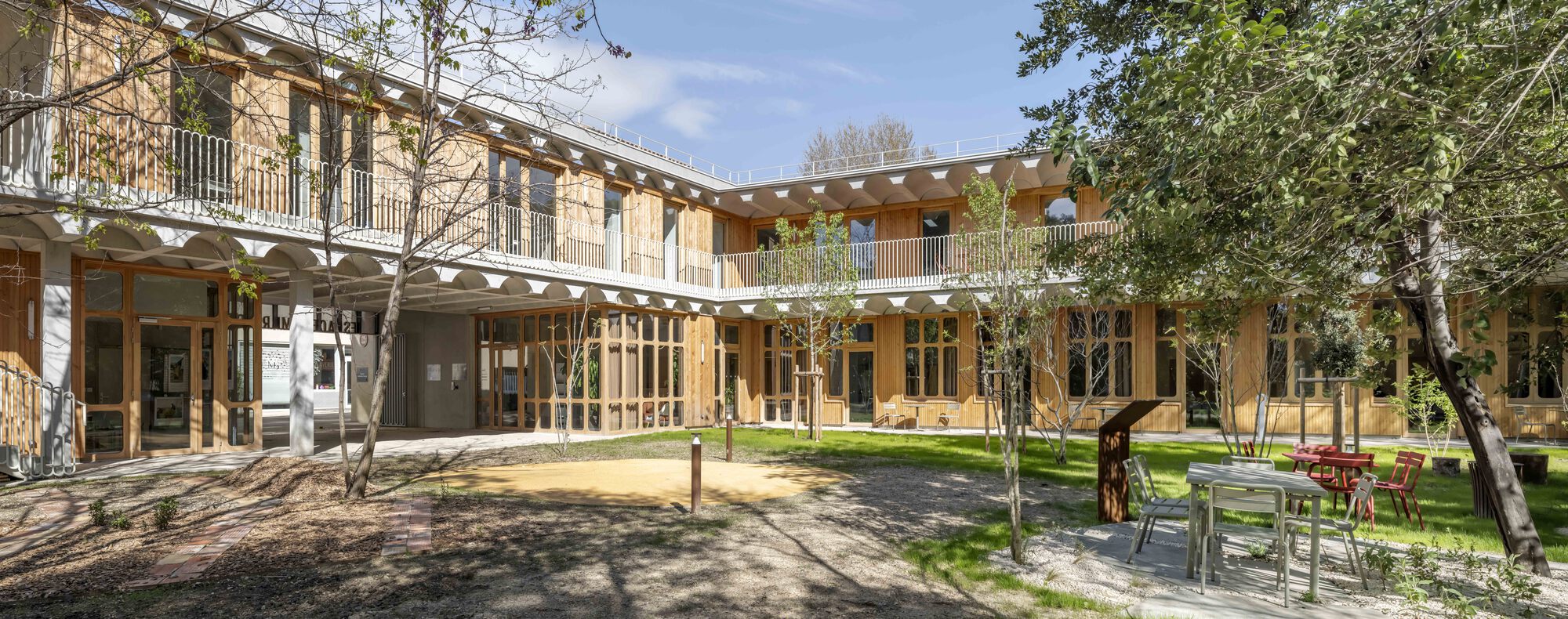 Wide view of the courtyard facades with timber-framed glazing, upper balcony and café tables beneath leafy trees